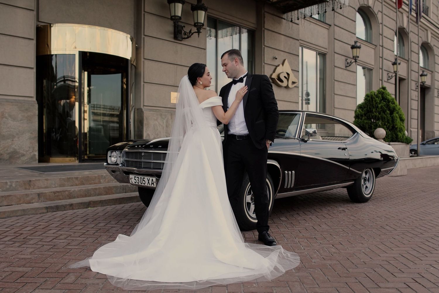 Bride and groom posing in front of a classic black vintage car outside an elegant hotel on their wedding day.