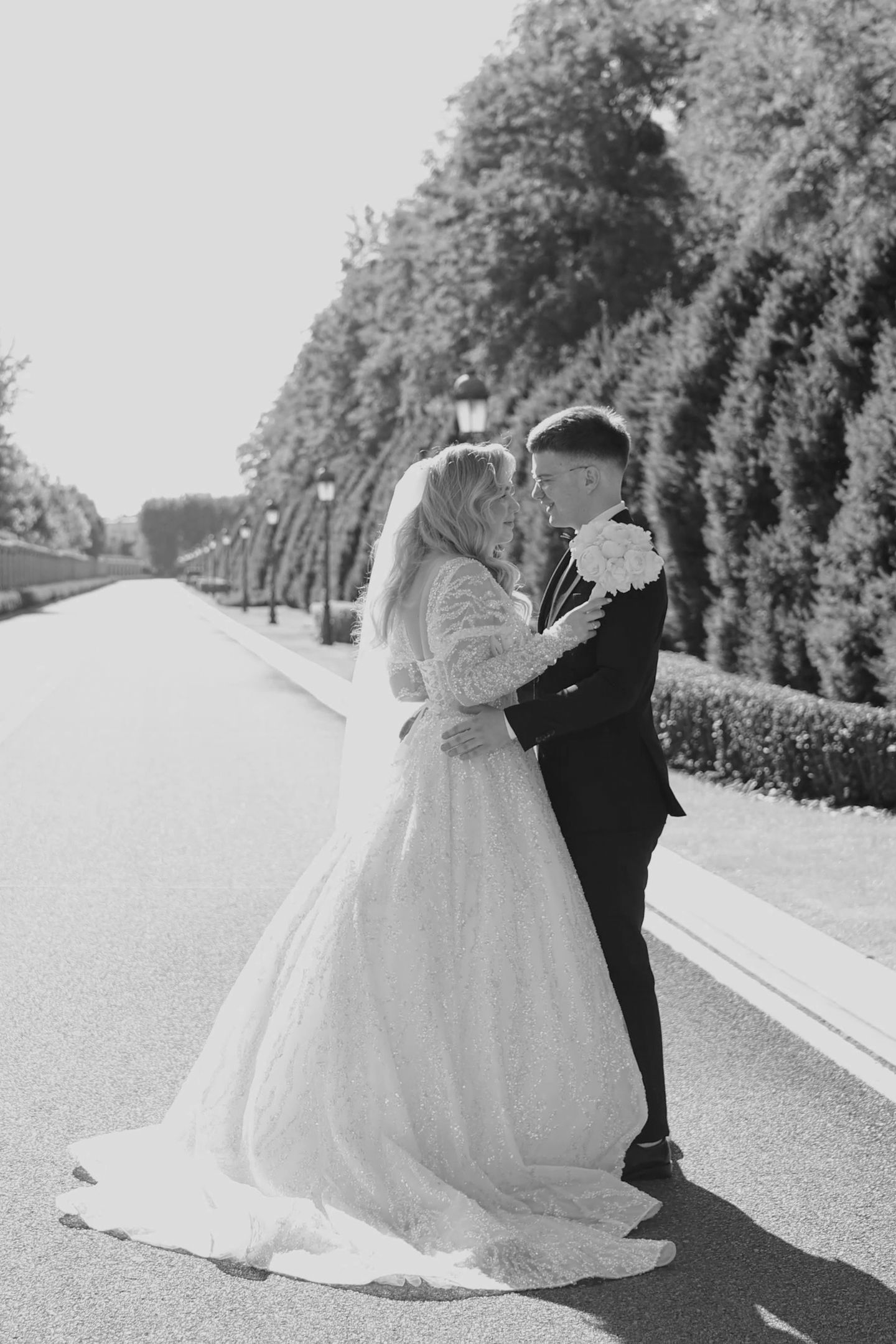 Black and white wedding photo of bride and groom embracing on a tree-lined path, bride holding bouquet.