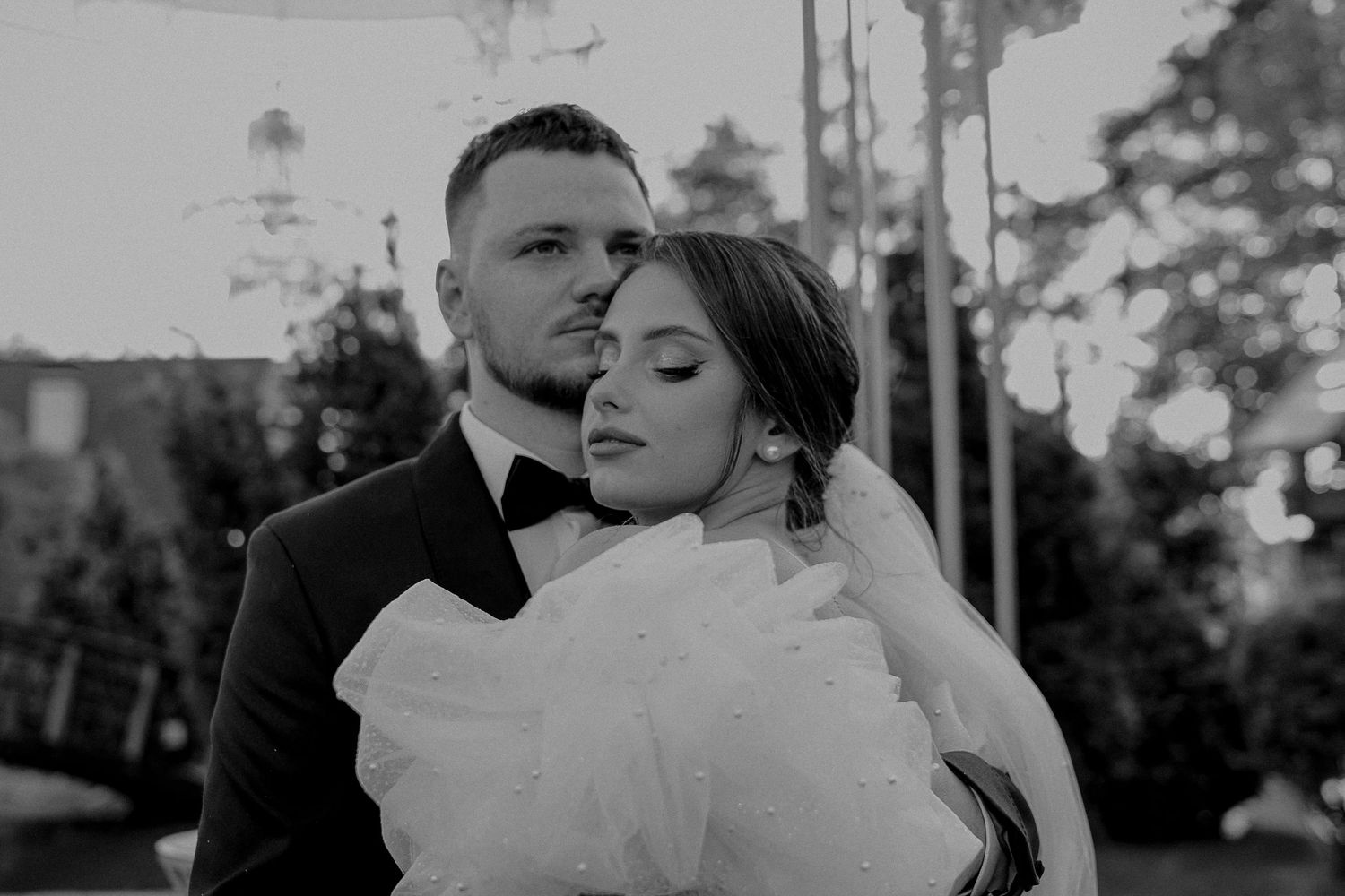Black and white wedding photo of bride and groom embracing outdoors, groom in tuxedo, bride in white gown.