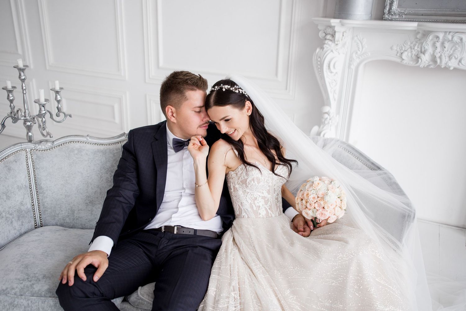A bride and groom sit closely together in an elegant white wedding photo studio.