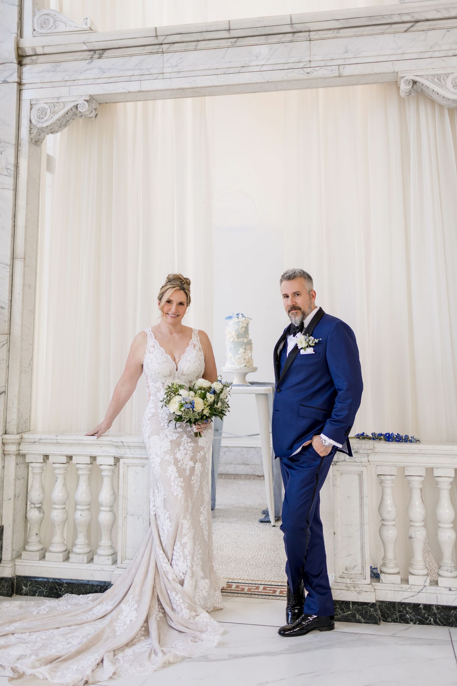 Bride in lace gown and groom in navy suit pose elegantly at a marble balustrade with white draping backdrop.