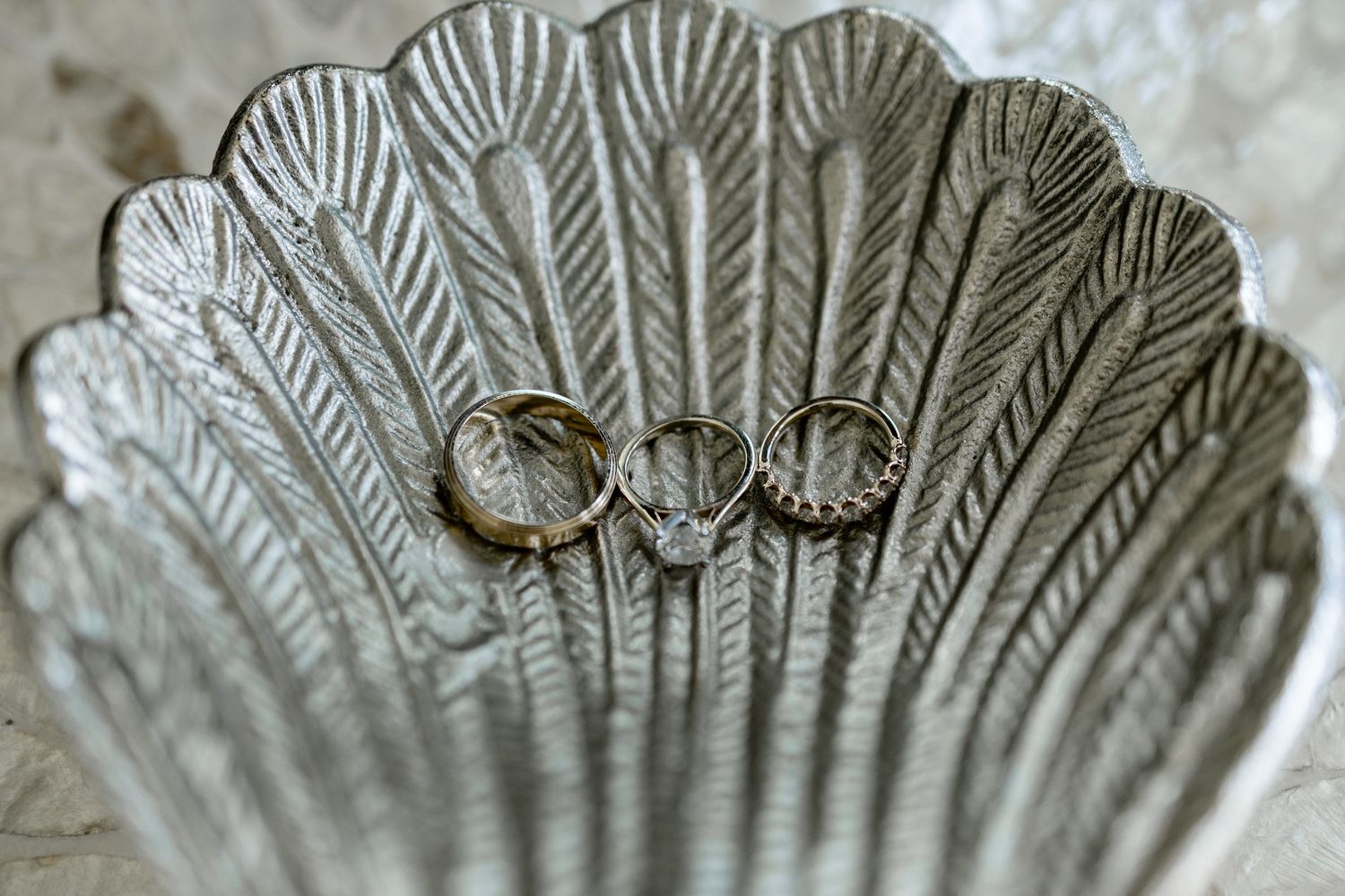 Three gold wedding rings resting in a decorative silver scallop shell dish on a marble surface.