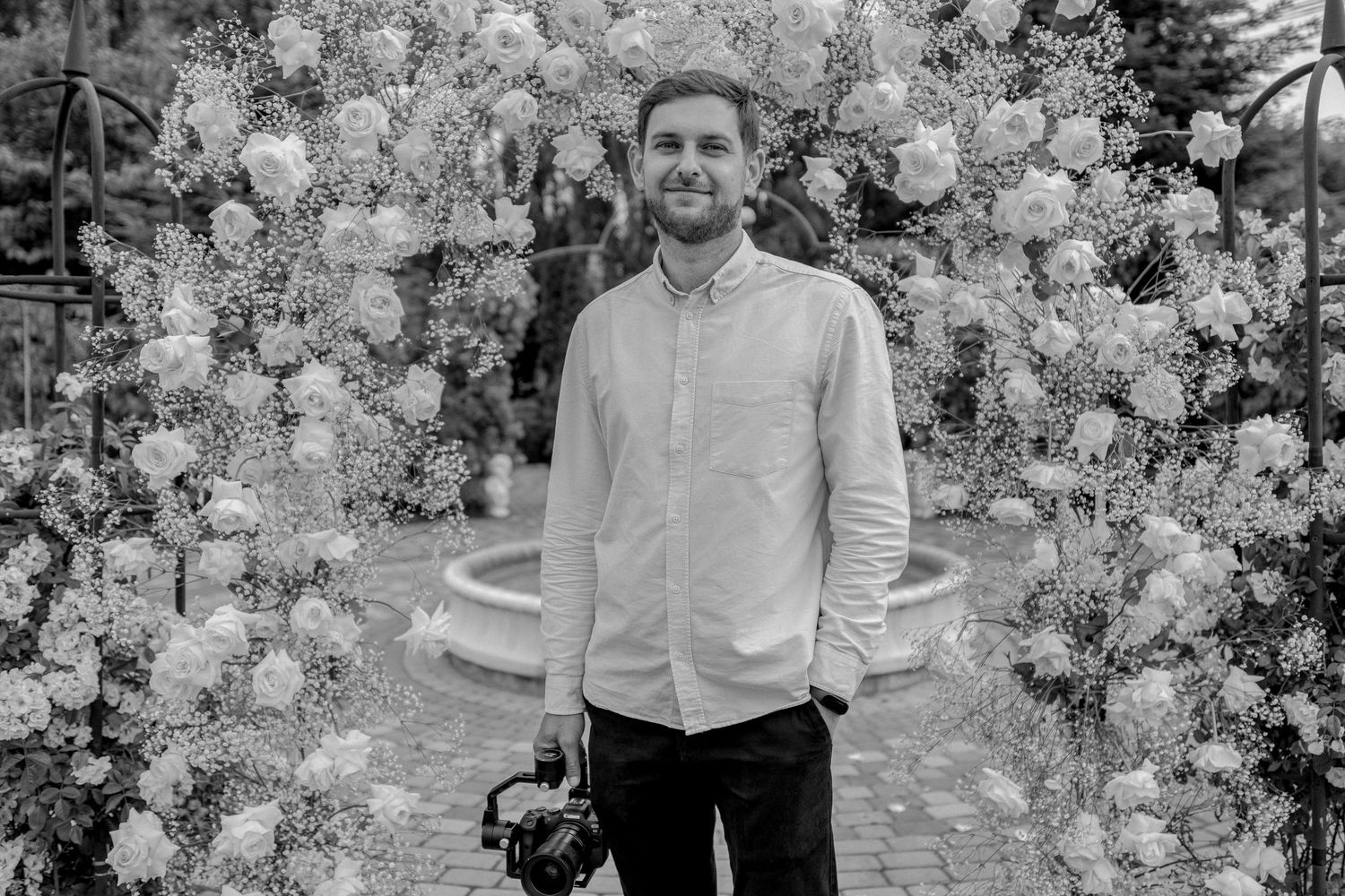 Black and white photo of a man holding a camera standing under a floral arch with a fountain backdrop.