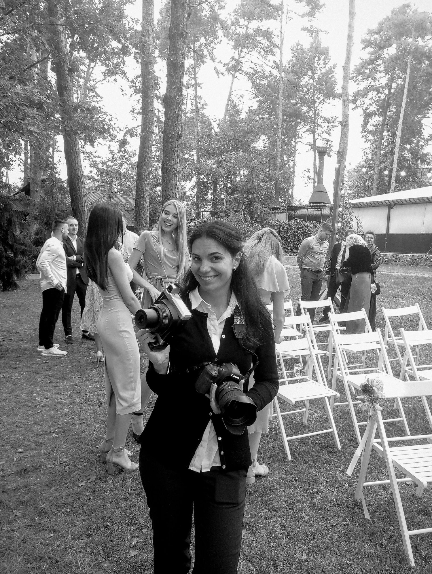 Black and white photo of a smiling female photographer holding two cameras at an outdoor wedding event.
