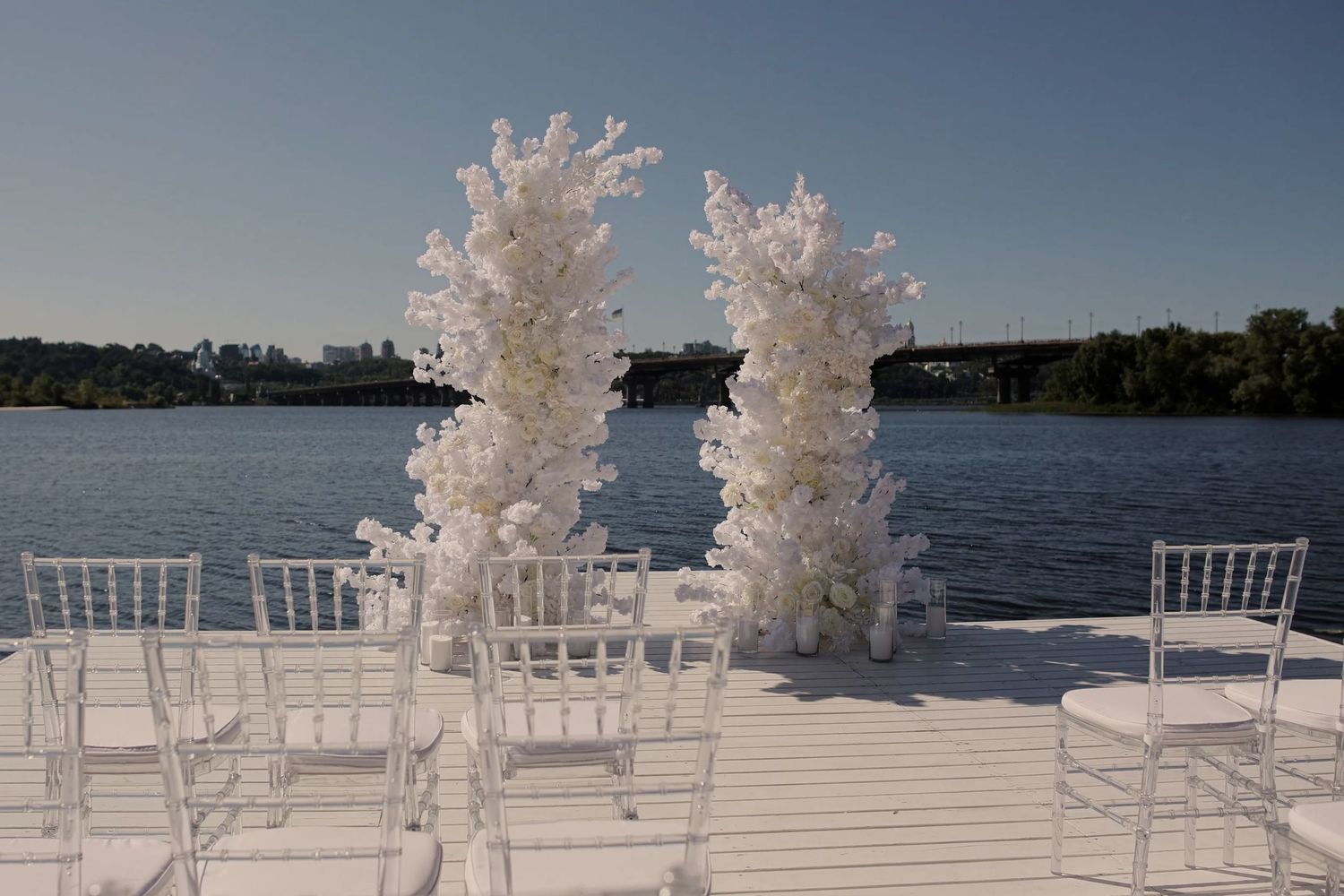 Elegant outdoor wedding ceremony setup on a waterfront dock with white floral arches and Chiavari chairs under a clear blue sky.