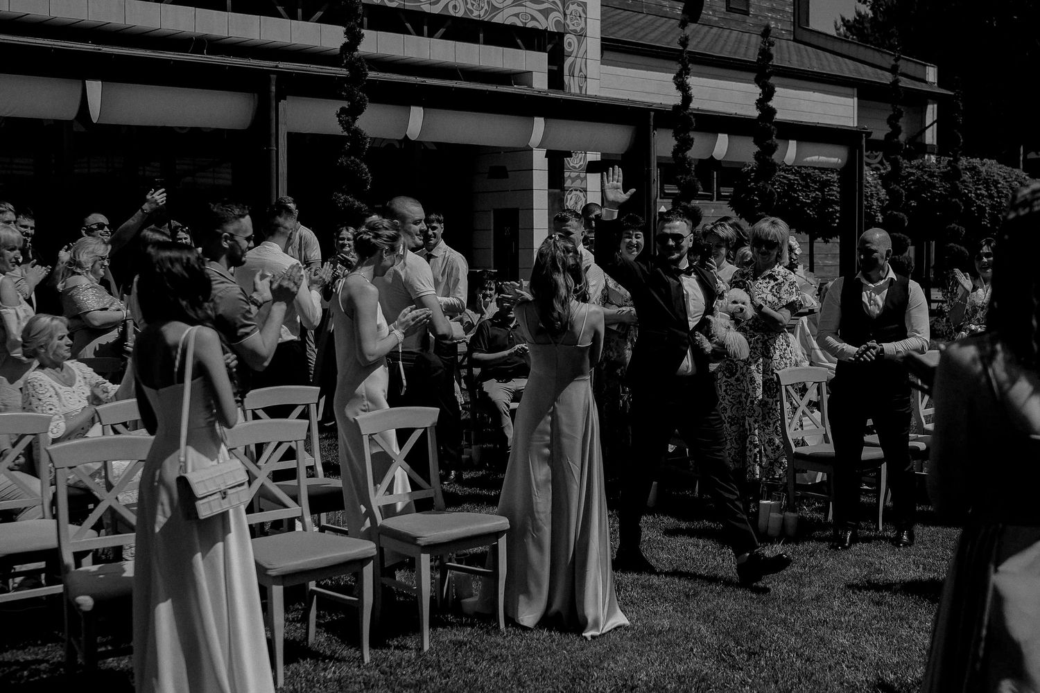 Black and white outdoor wedding ceremony with guests gathered on lawn beside elegant venue building.