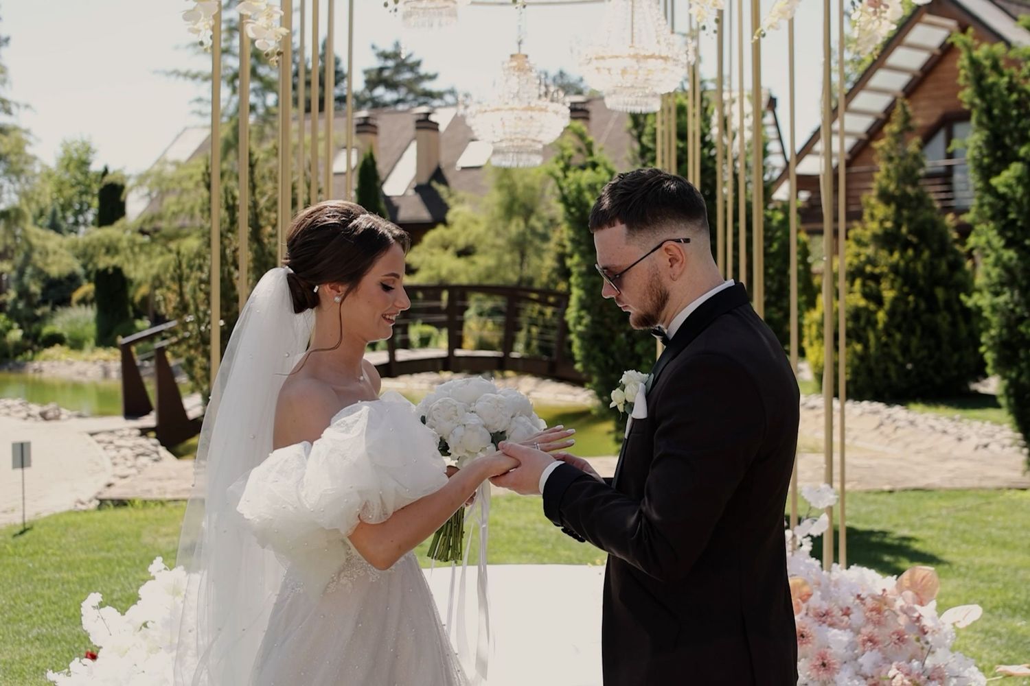 Bride and groom exchanging rings during outdoor wedding ceremony with floral arch and lush green garden backdrop.