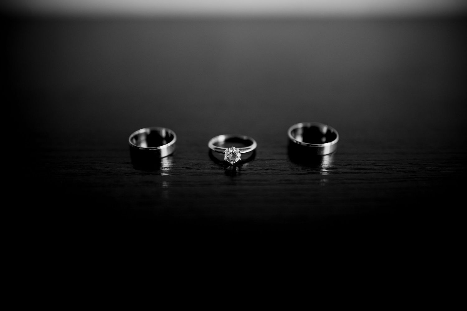 Three silver rings on dark surface, center diamond engagement ring flanked by two wedding bands, black and white photography.