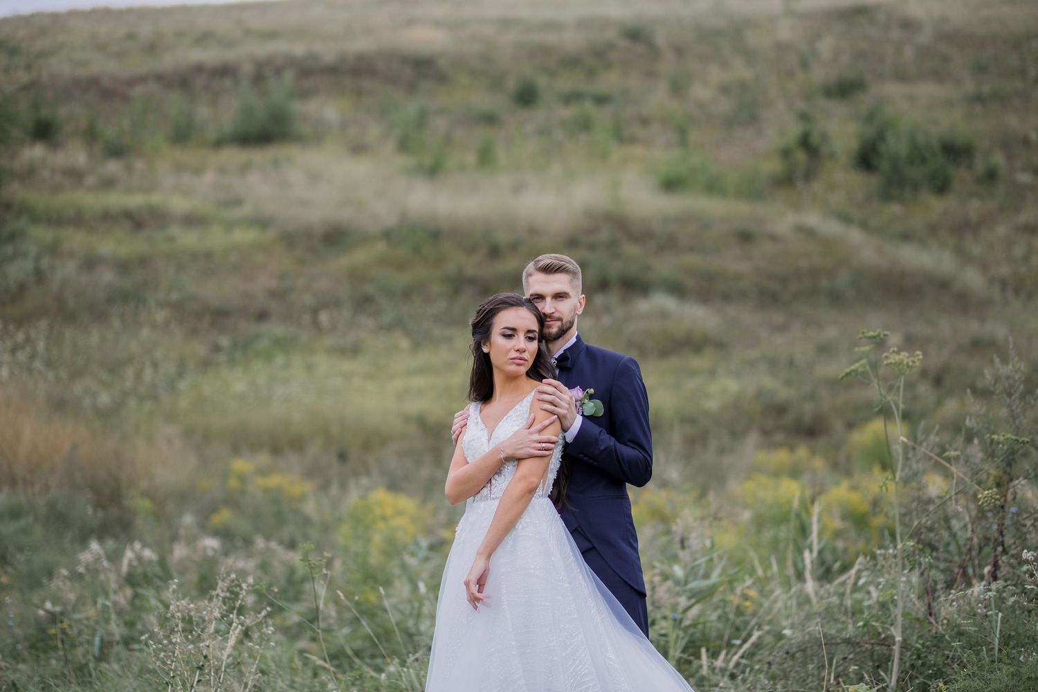 Romantic wedding couple embracing in a green meadow, bride in white gown and groom in navy suit holding flowers.