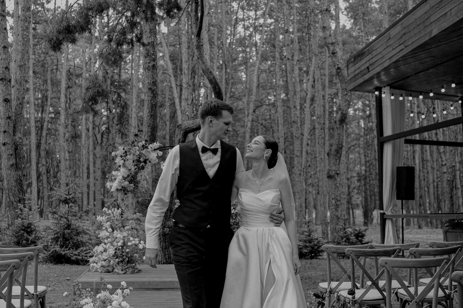 Black and white photo of a bride and groom walking together in a forest wedding venue with string lights.