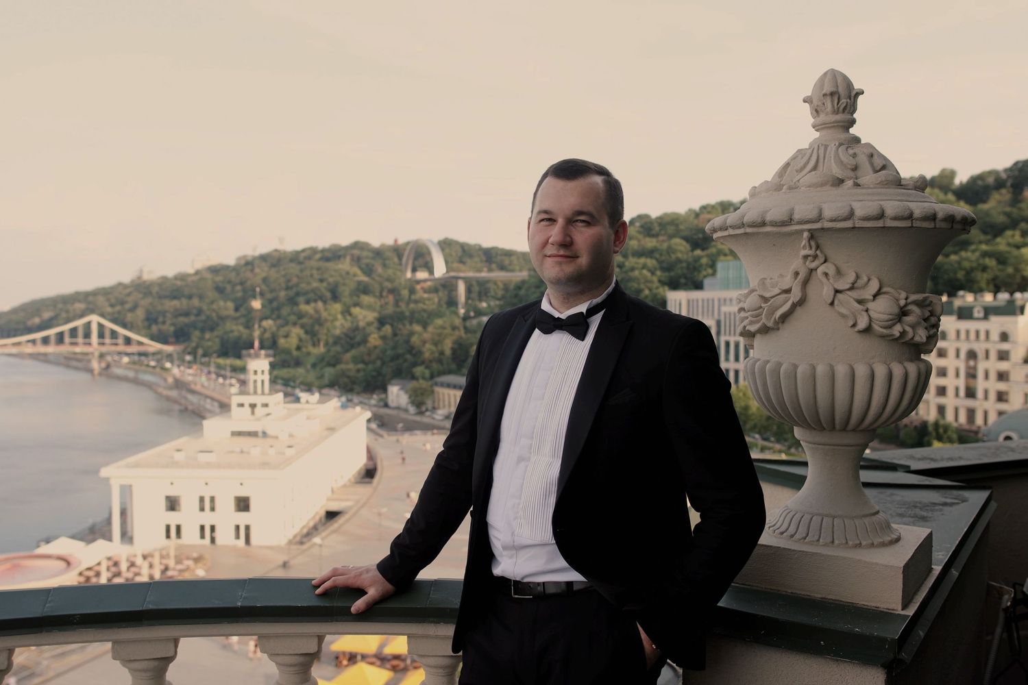 Elegant man in tuxedo posing on balcony with scenic river and green hills backdrop.