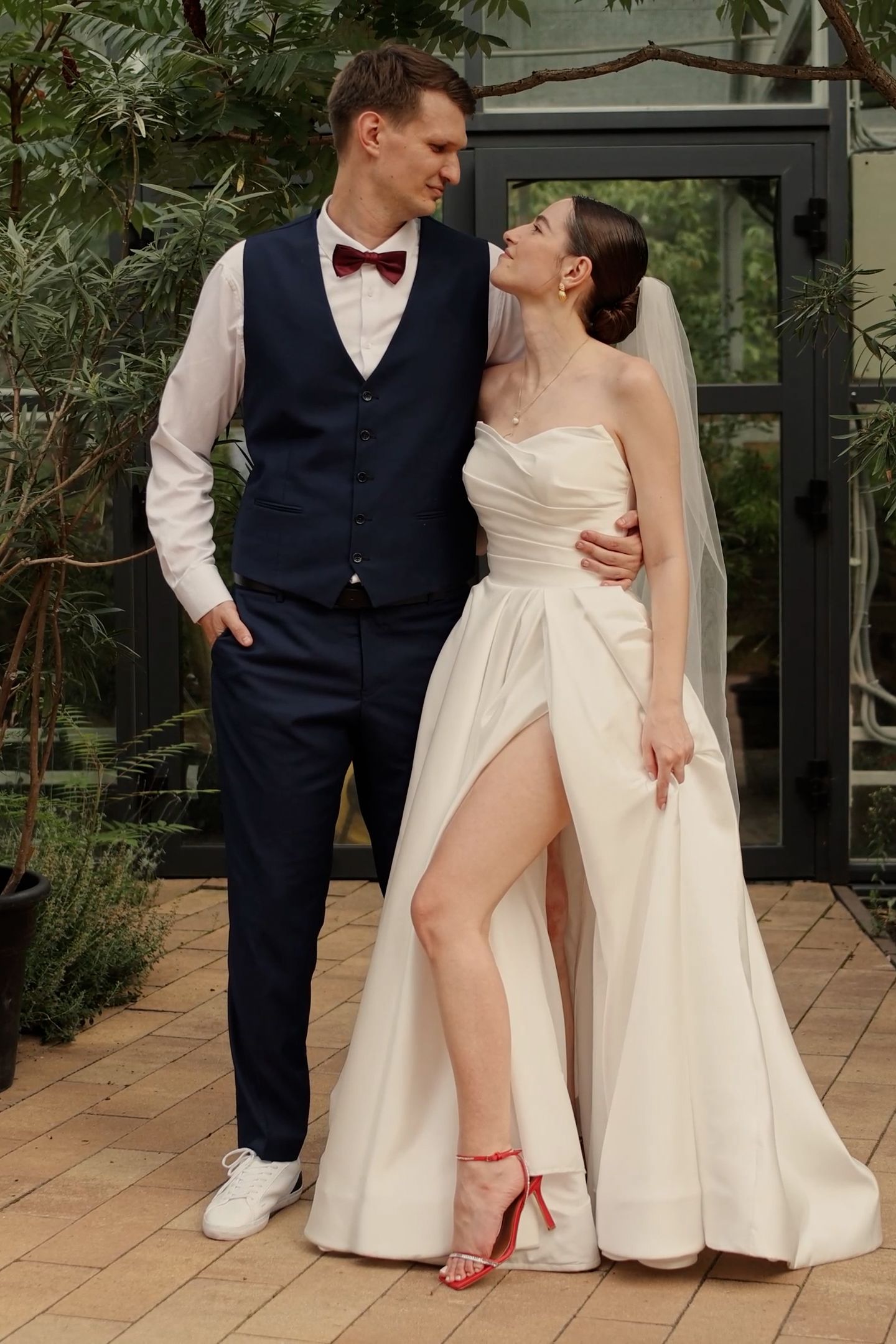 Elegant bride in white strapless gown with slit and groom in navy vest with burgundy bow tie posing outdoors.