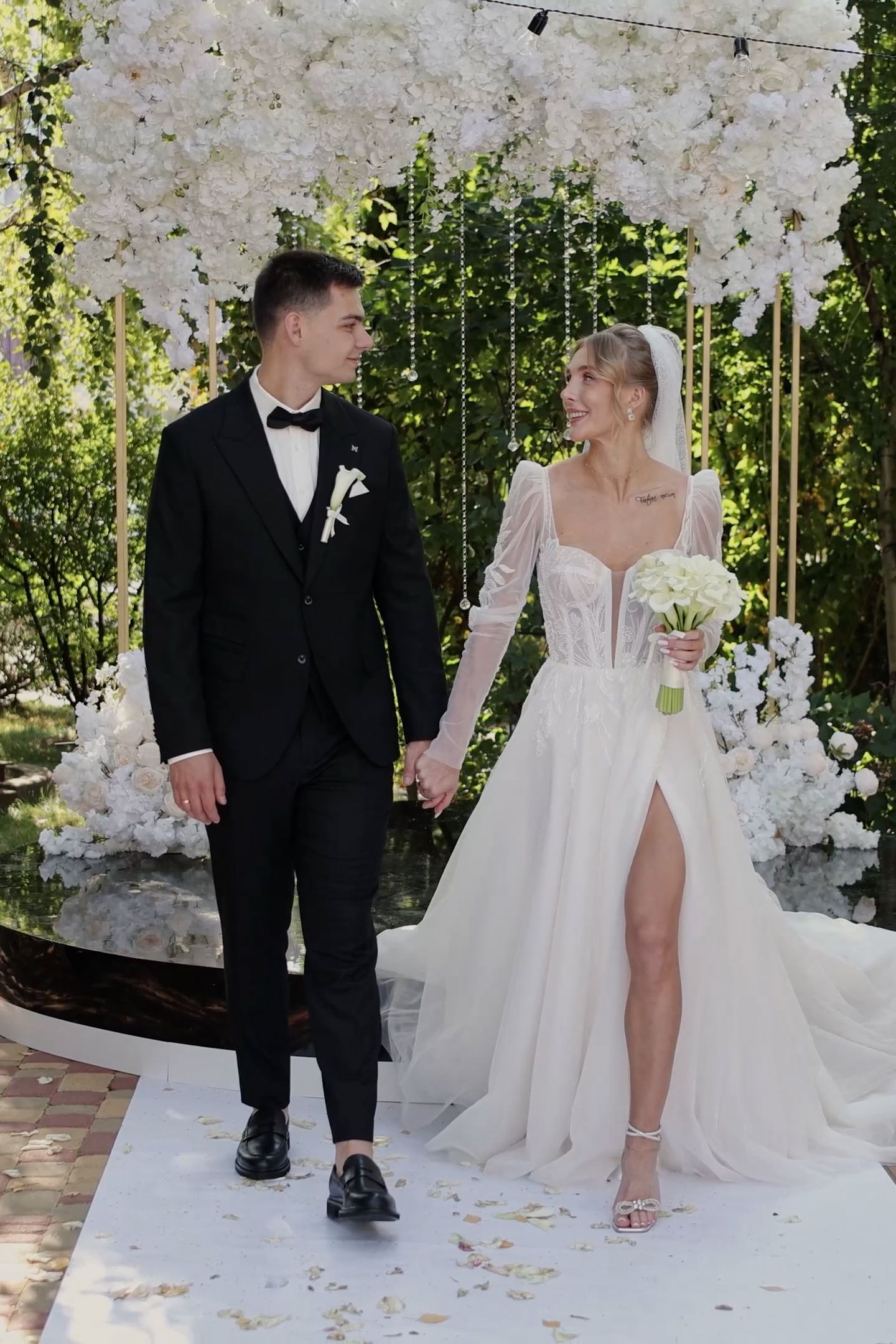 Bride and groom holding hands at outdoor wedding ceremony with white floral arch backdrop.