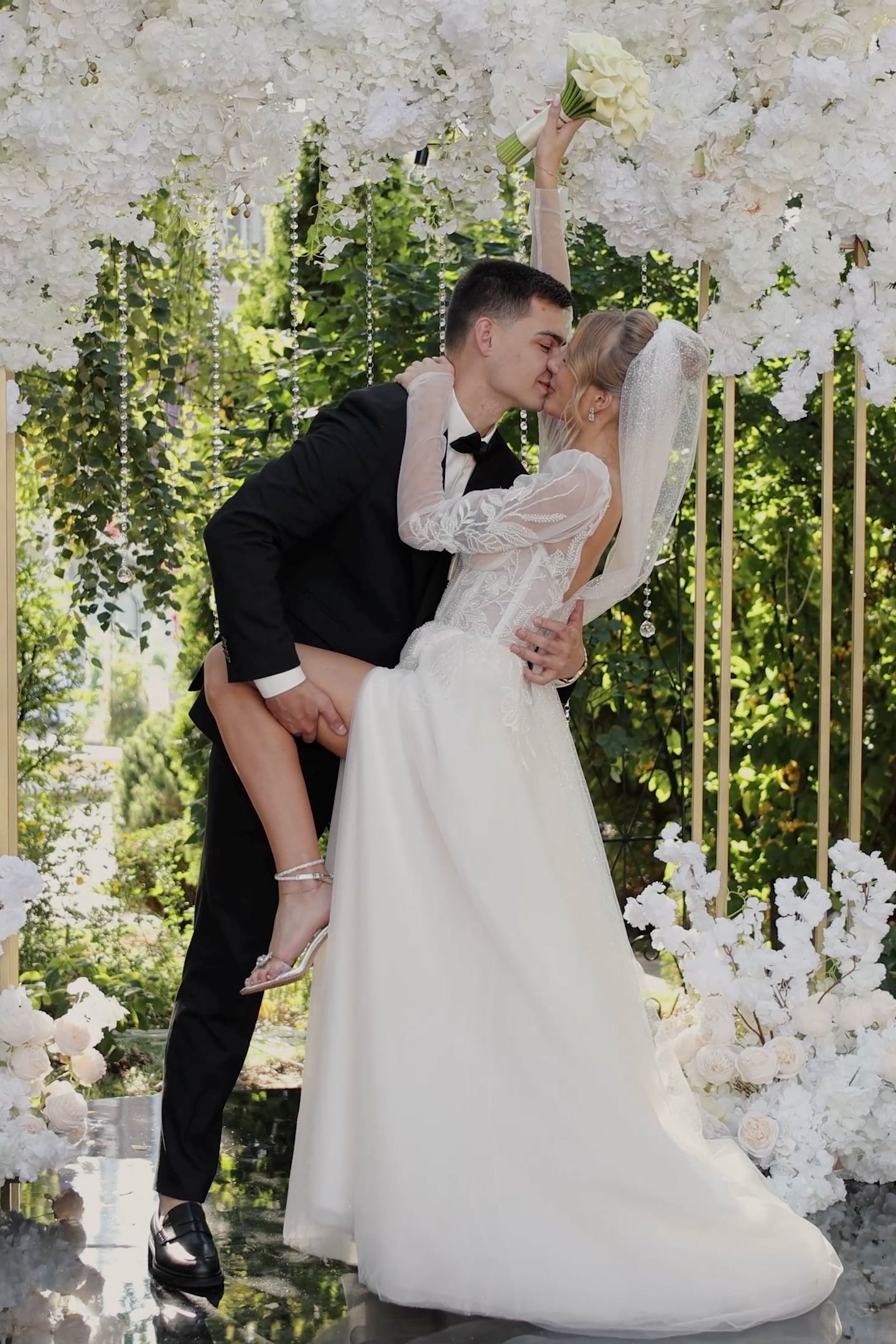 Bride and groom share a romantic dip kiss at outdoor wedding ceremony surrounded by white floral arch decorations.