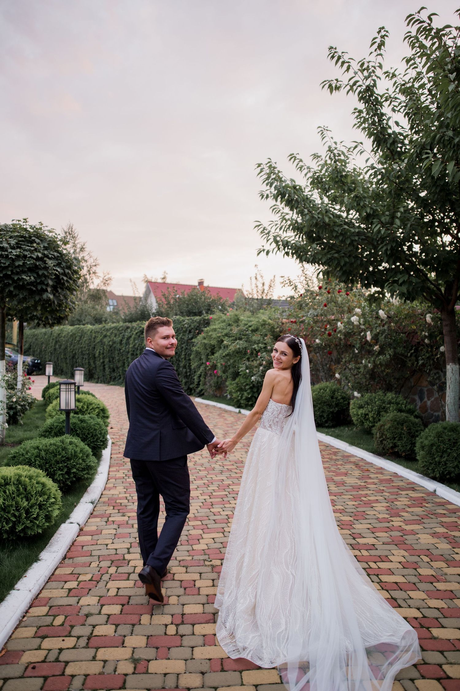 Bride and groom holding hands walking on brick path in lush garden, bride in elegant white gown with veil.