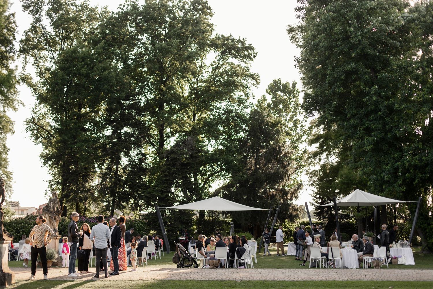 Outdoor garden wedding reception with guests mingling under white canopies surrounded by tall green trees.