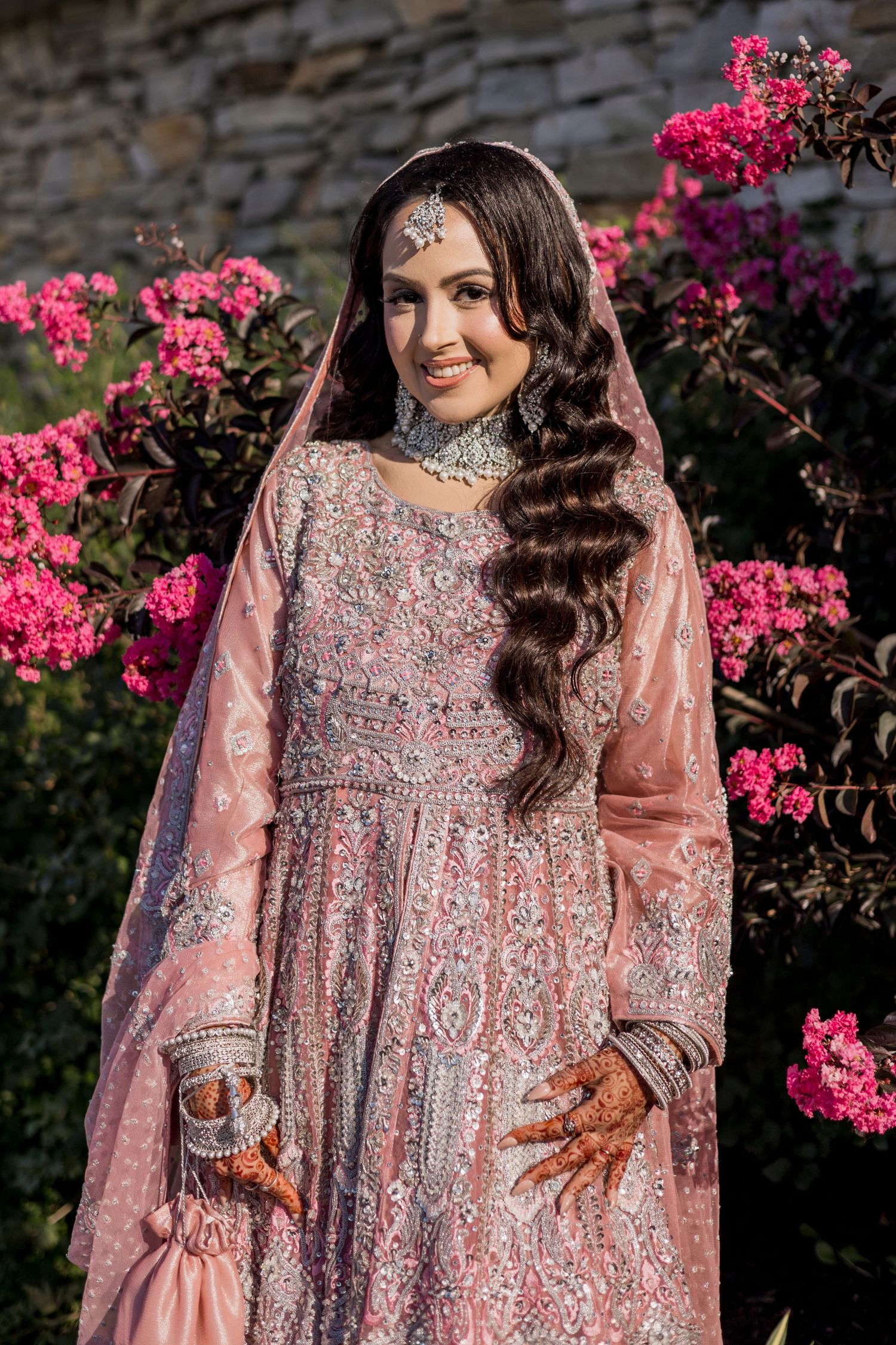 A bride in an embroidered pink lehenga with dupatta and henna, posing among vibrant pink bougainvillea flowers.