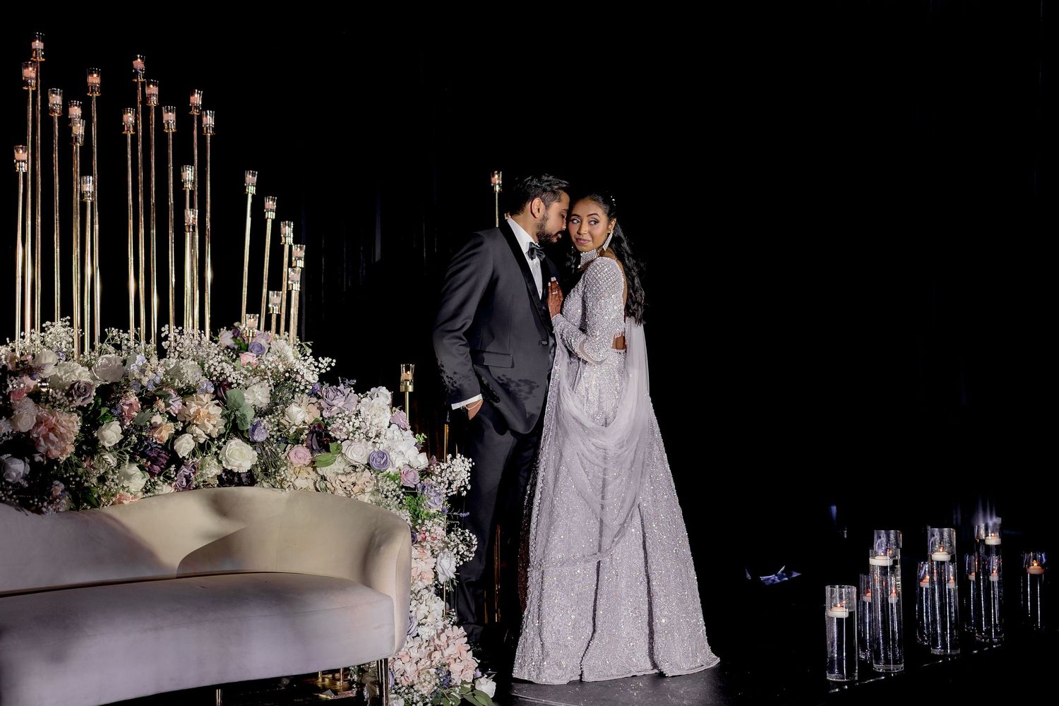 Elegant wedding couple sharing a romantic moment on a dark stage surrounded by floral arrangements and tall gold candelabras.