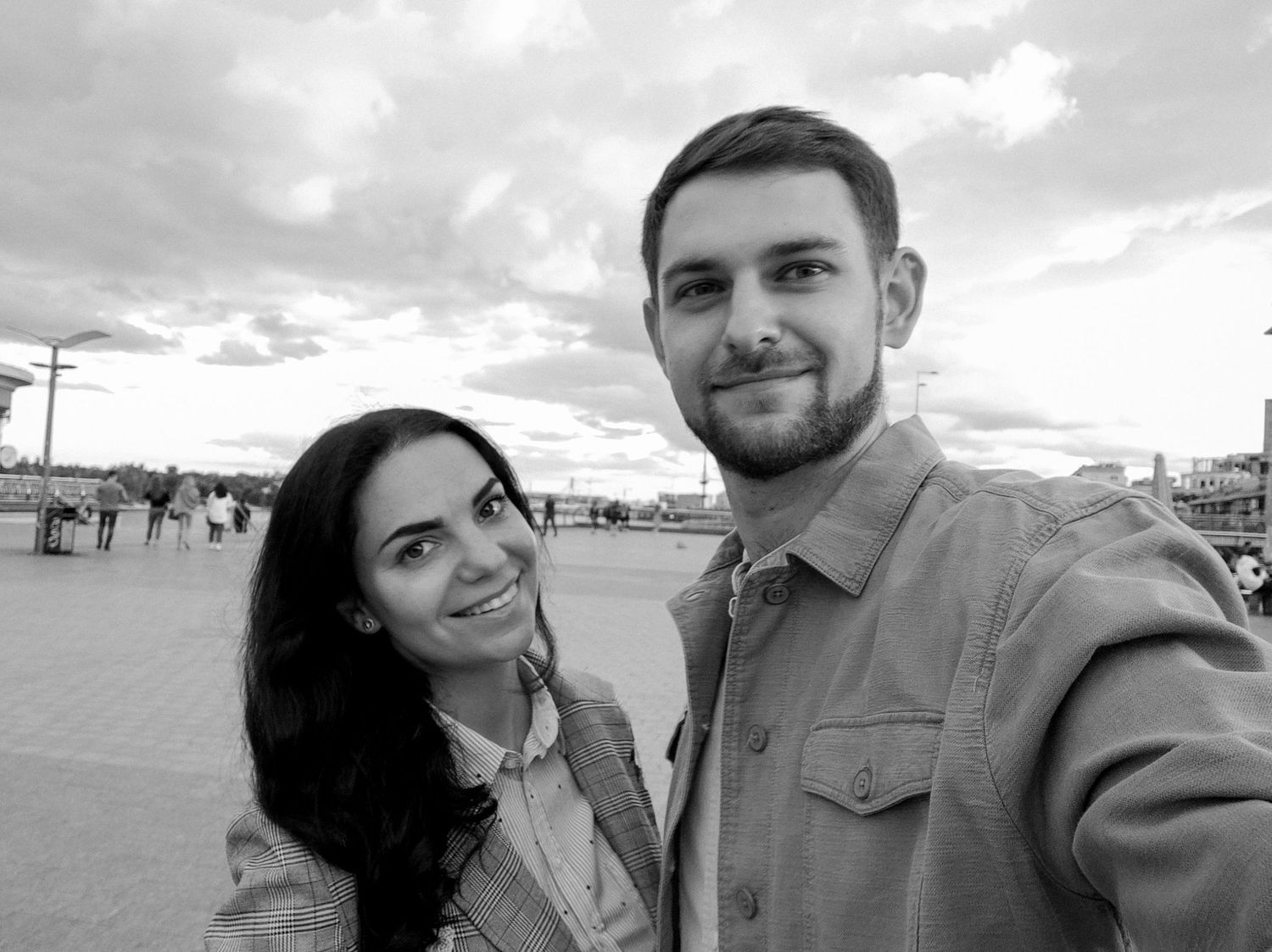 Black and white selfie of a smiling young couple outdoors with a cloudy sky backdrop.