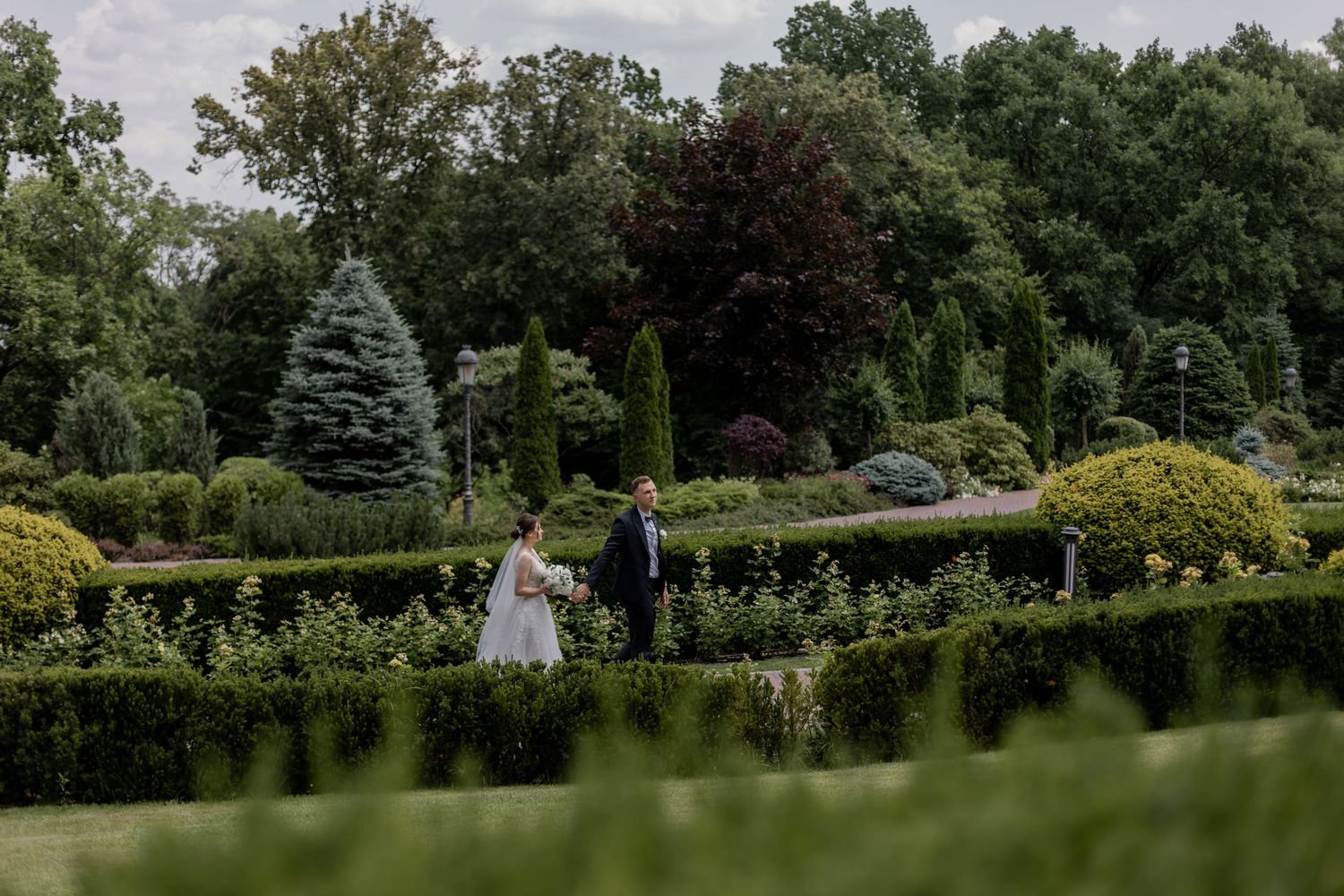 A bride and groom walk hand in hand through a lush formal garden with manicured hedges and tall trees.