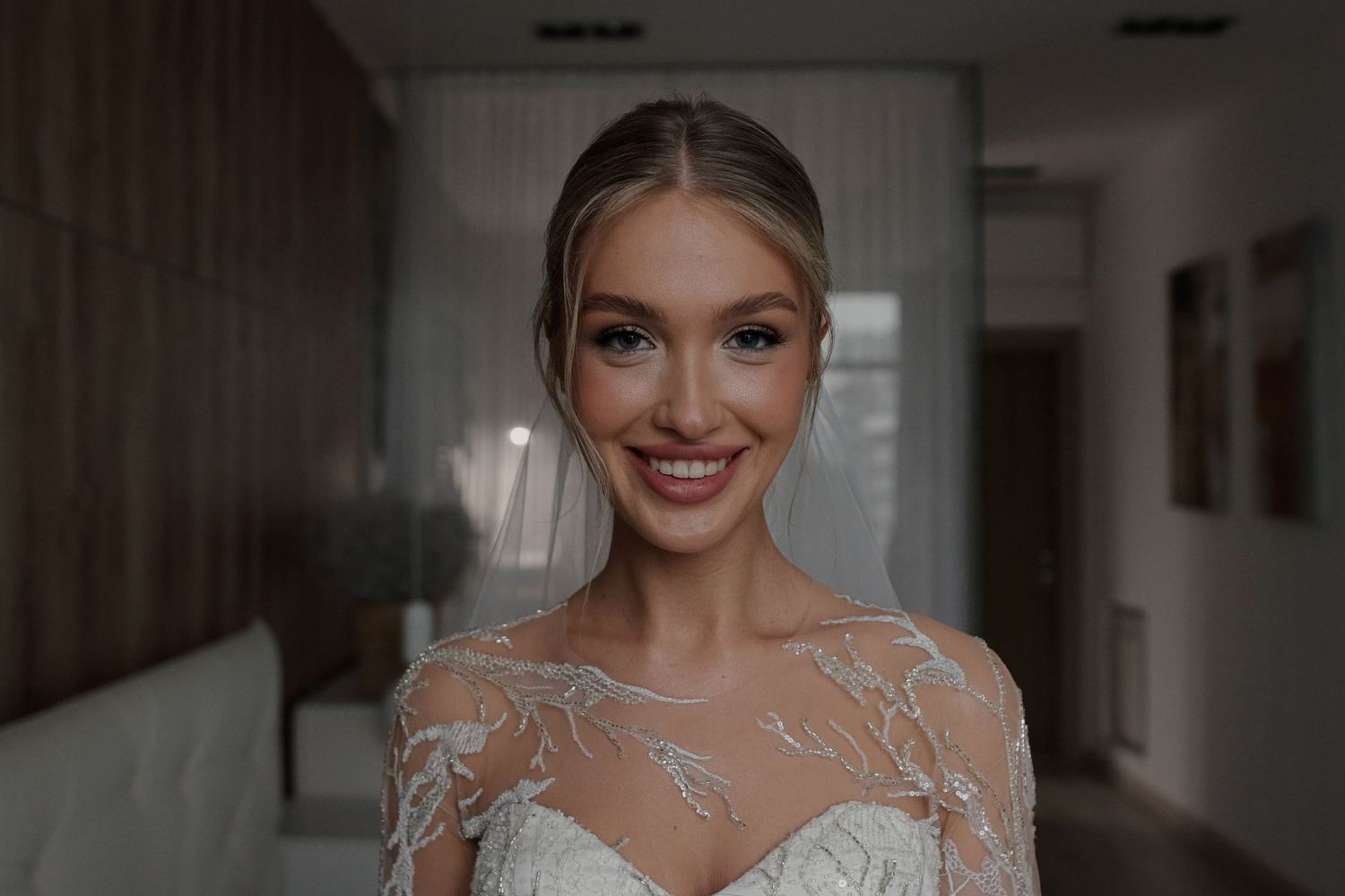 Smiling bride in elegant lace wedding gown with sheer sleeves, wearing updo hairstyle and natural makeup indoors.