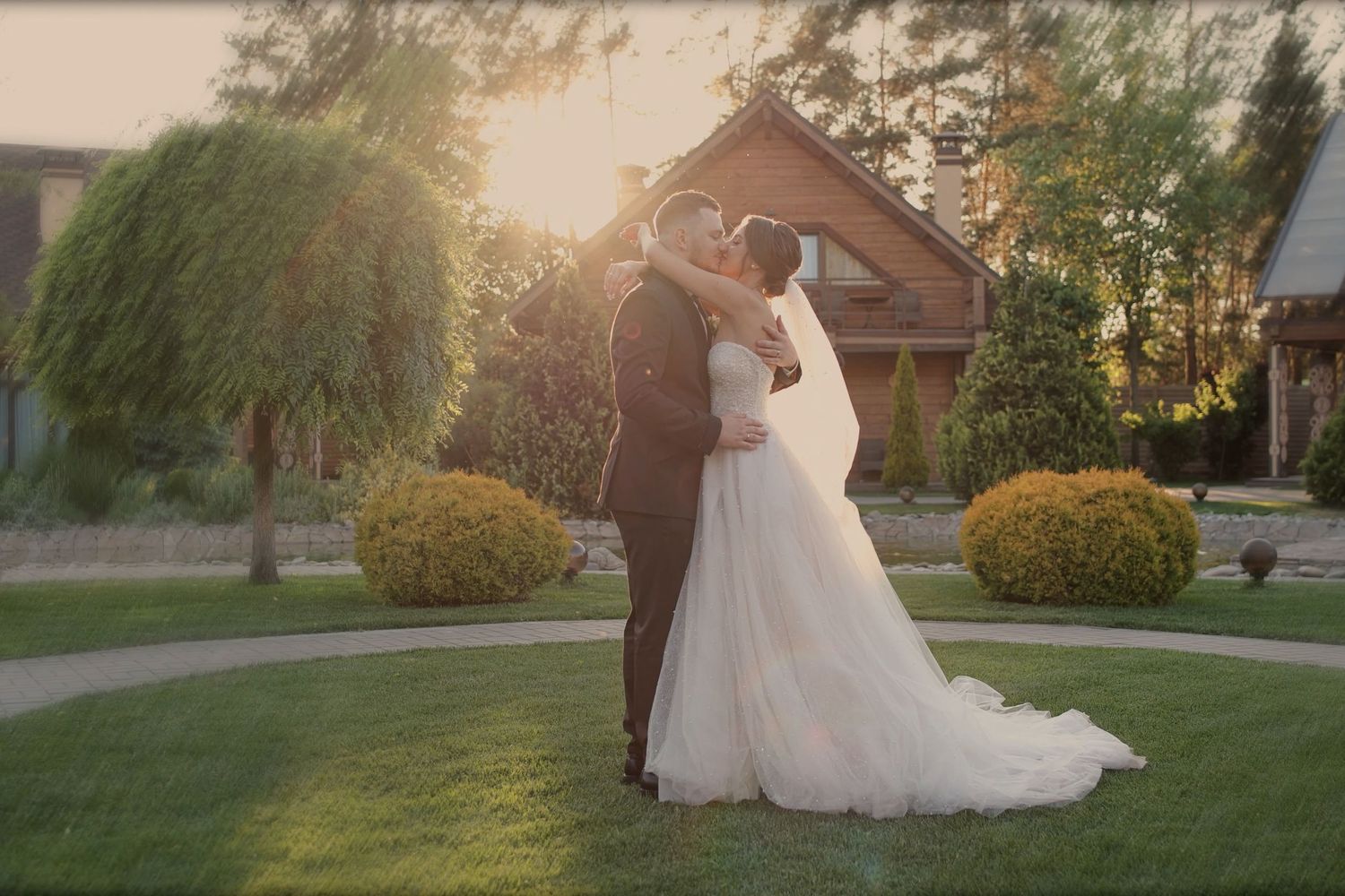 Bride and groom sharing a romantic kiss at golden hour in a lush garden with warm sunlight glowing behind them.