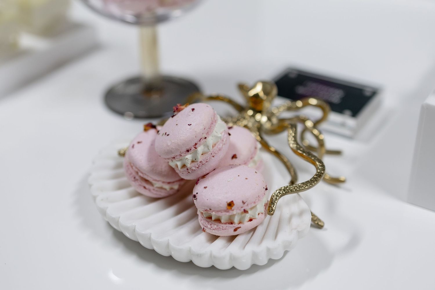 Pink macarons with sprinkles on a white shell dish, decorated with gold jewelry on a white surface.