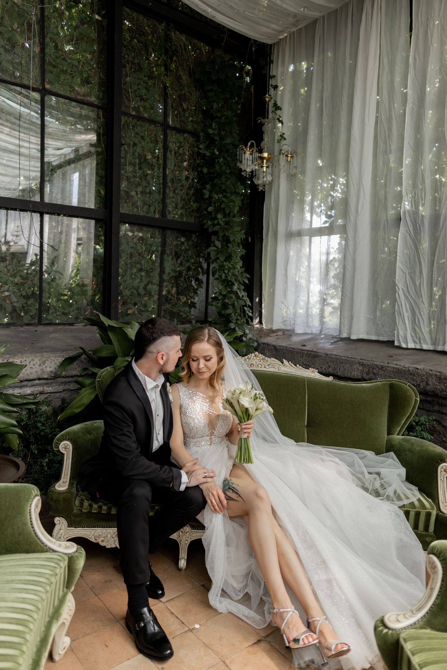 Bride and groom sitting on green velvet sofa in elegant greenhouse venue, bride holding bouquet in lace wedding gown.
