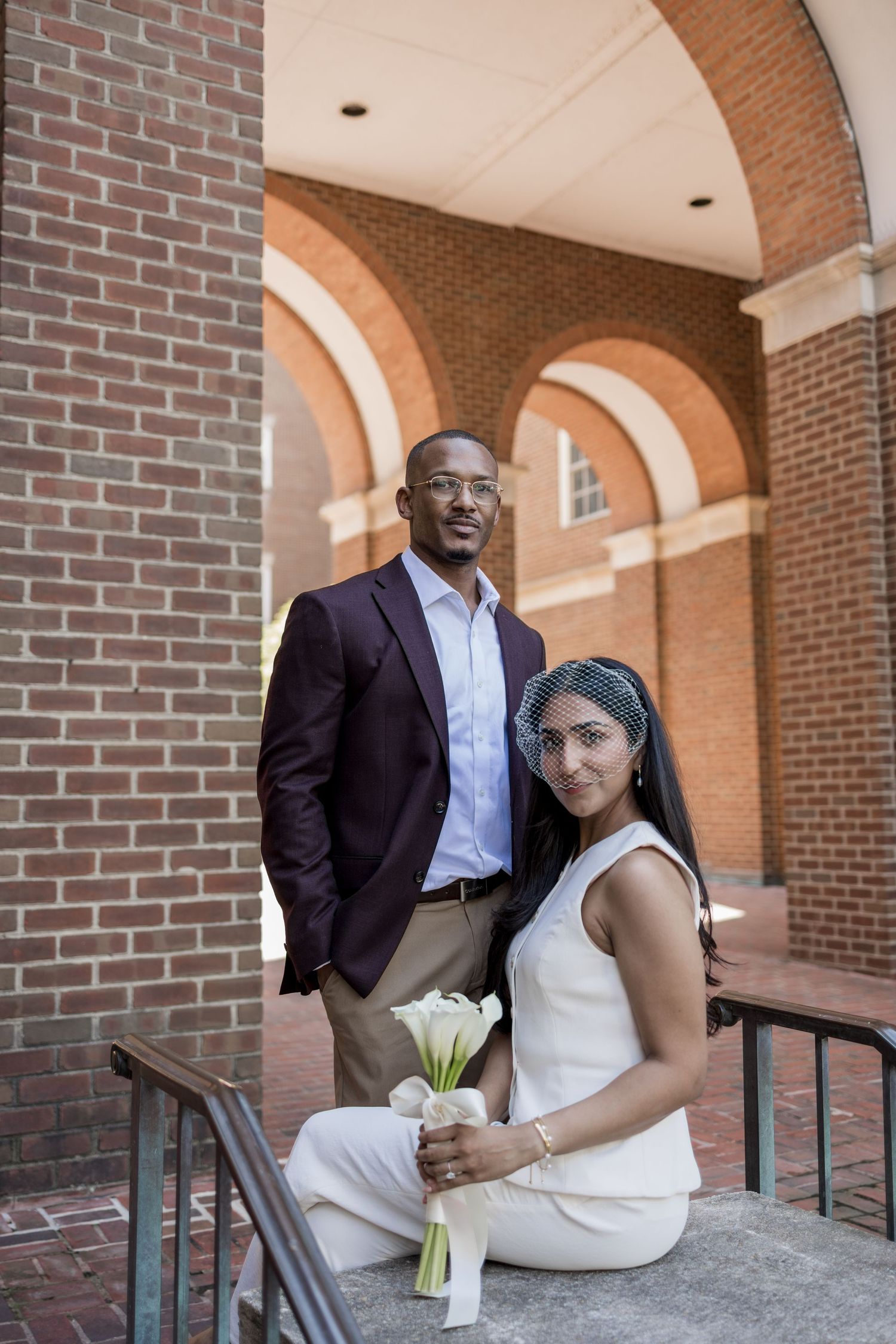 Couple posing at brick building with arches, woman in white jumpsuit holding calla lilies, man in burgundy blazer.