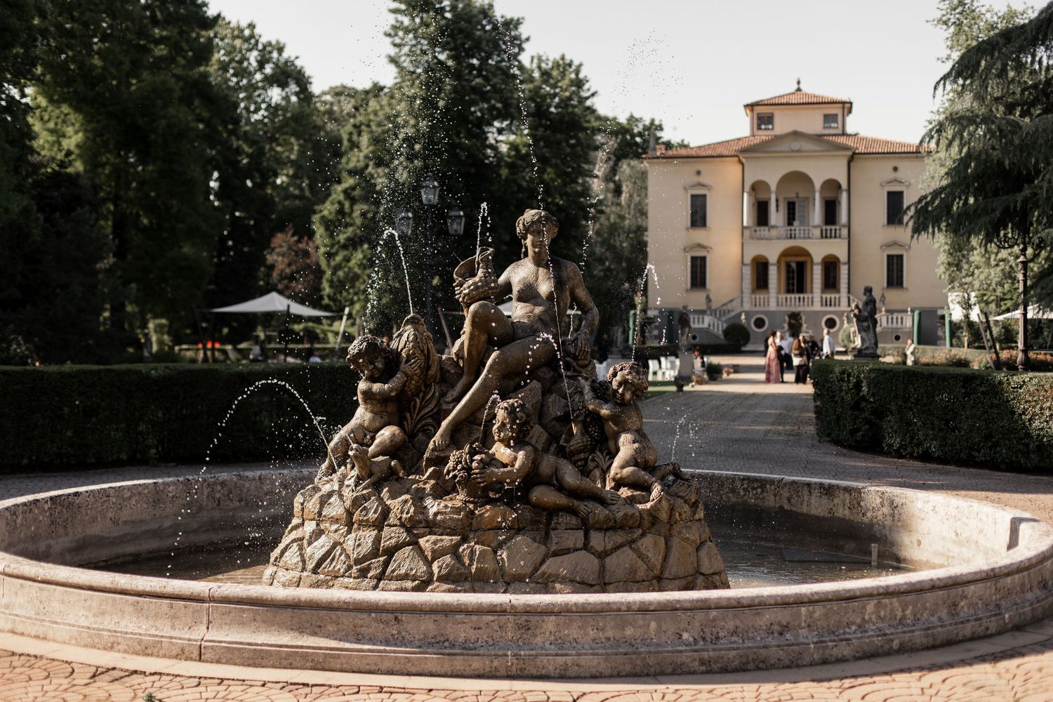 Bronze fountain sculpture of a goddess with cherubs in an Italian garden plaza with a historic villa backdrop.