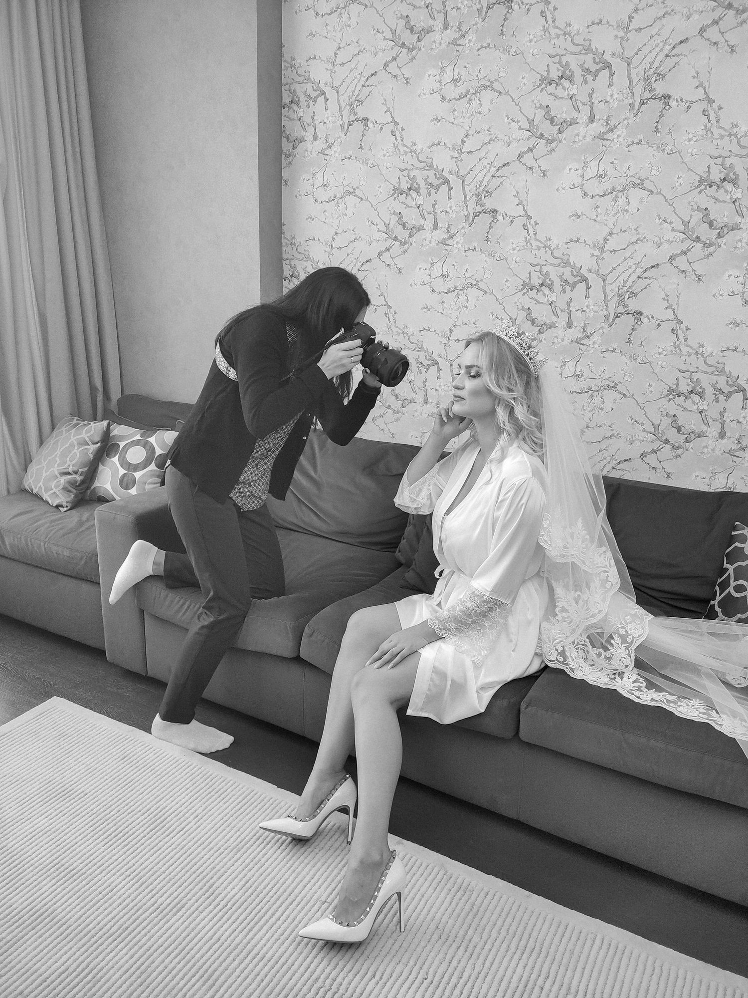 Black and white photo of a photographer capturing a bride in a satin robe and veil seated on a sofa.