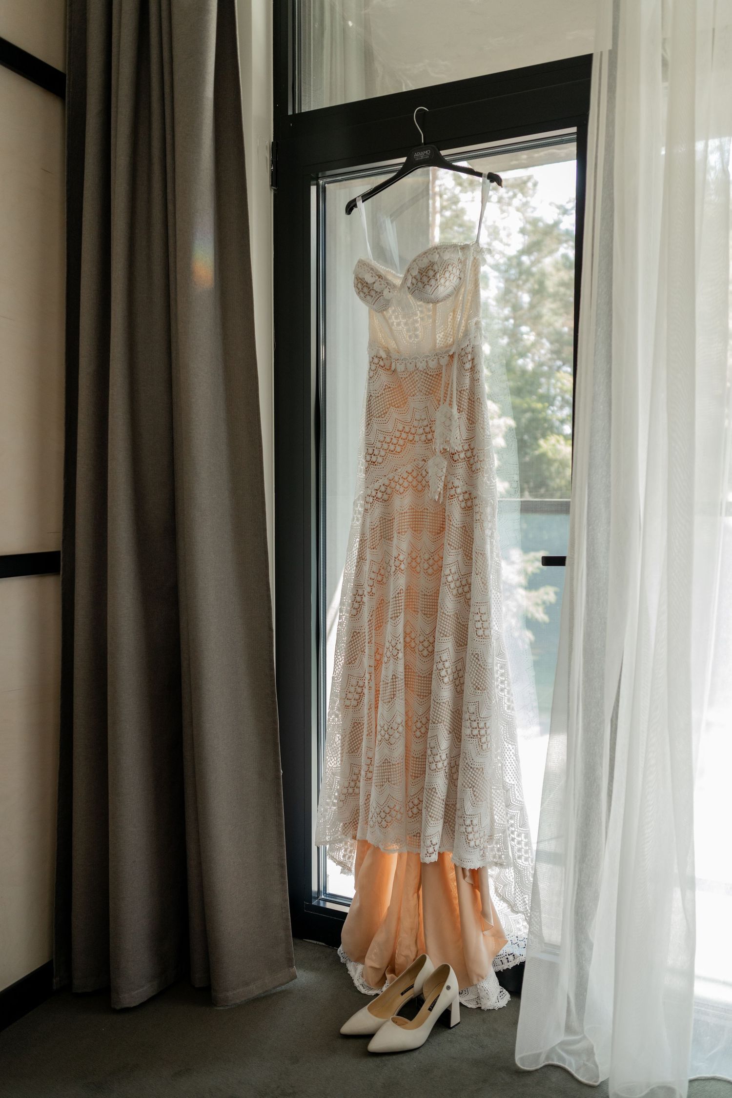 Wedding dress and shoes displayed hanging by window with sheer curtains and natural lighting.