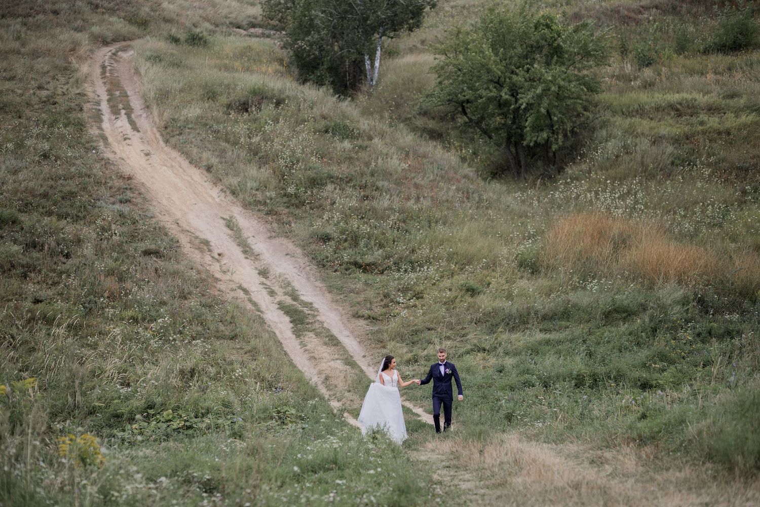 A bride and groom walk hand in hand along a winding dirt path through a lush green countryside field.