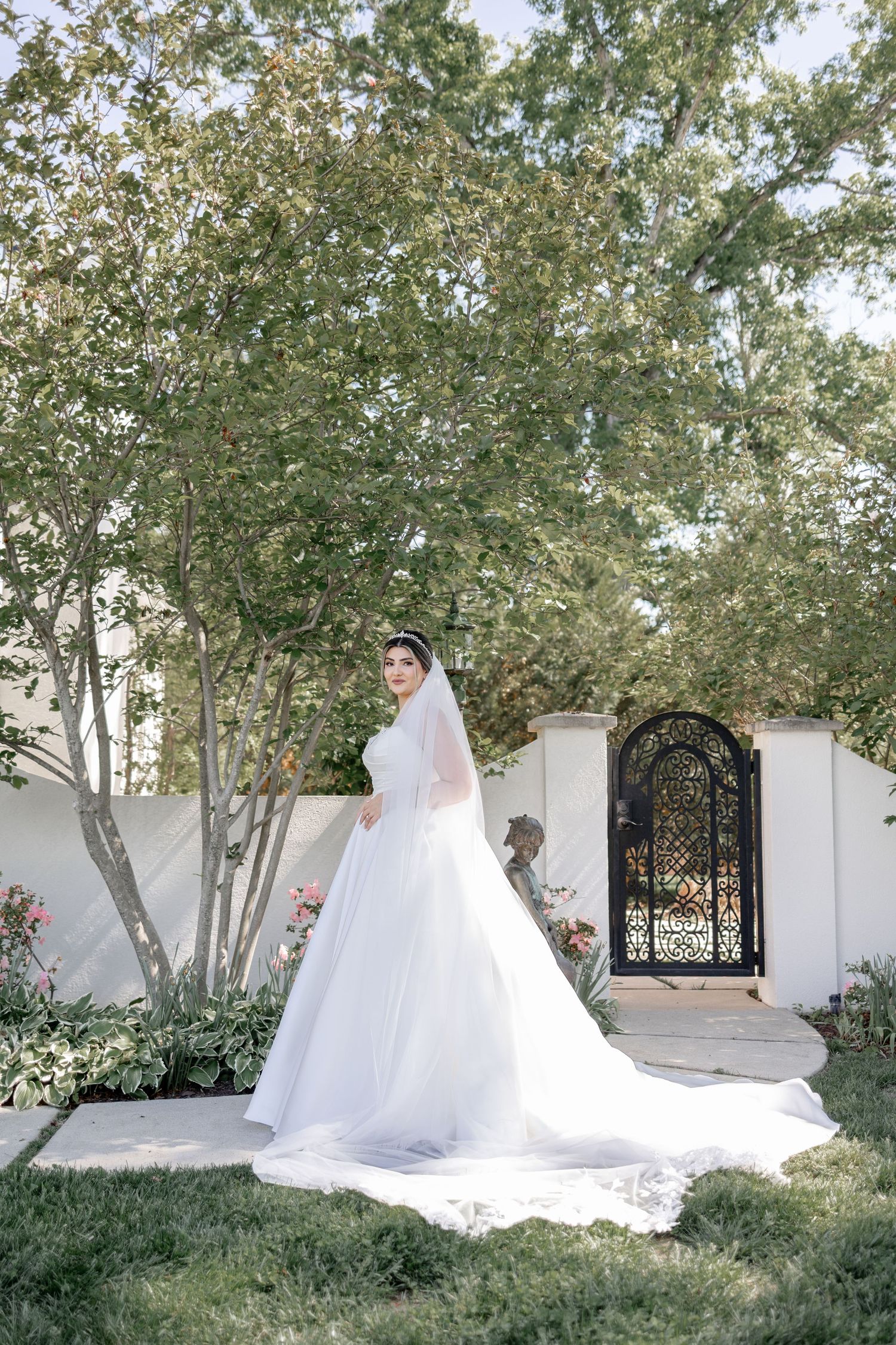 Bride in elegant white ball gown and long veil poses in lush garden with ornate iron gate backdrop.