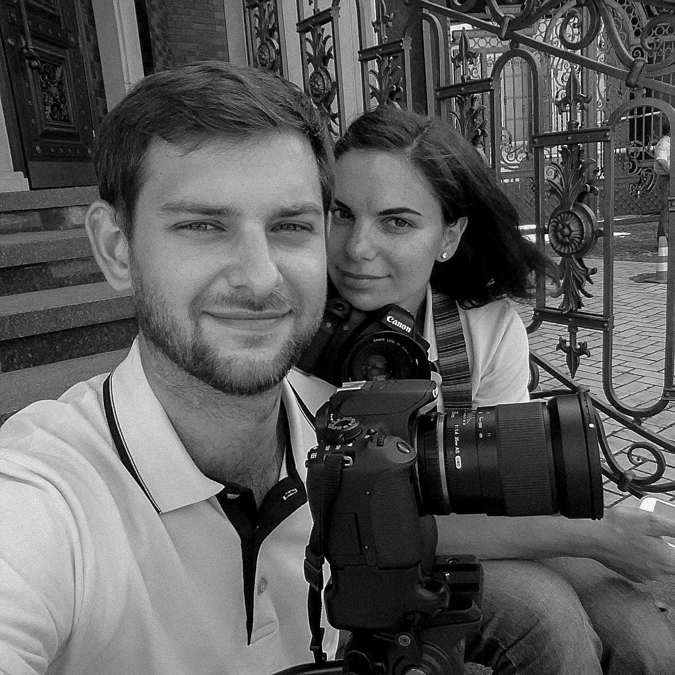 Black and white photo of a young couple taking a selfie with a DSLR camera outdoors near ornate iron gates.