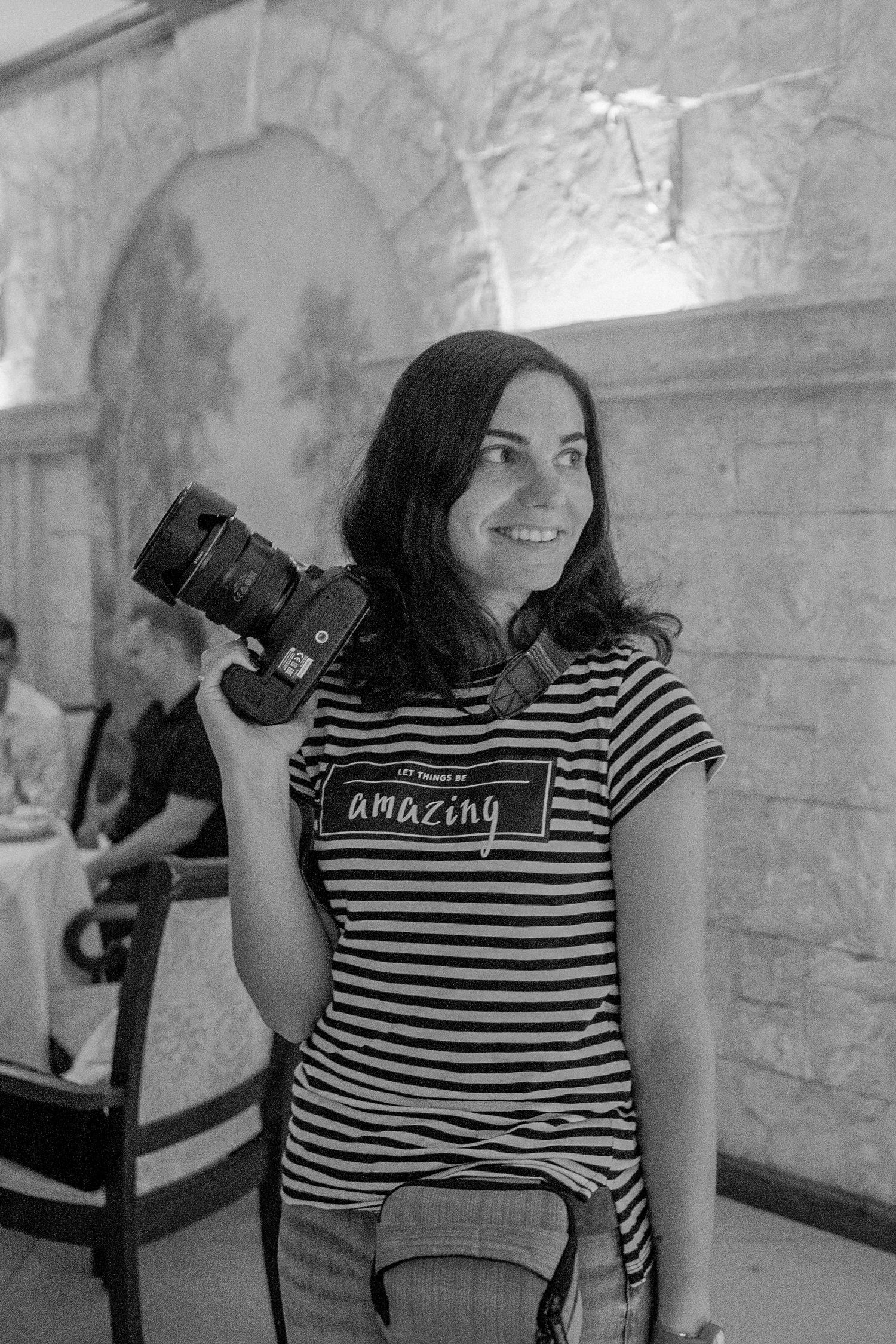 Black and white photo of a smiling woman holding a camera, wearing a striped t-shirt that reads amazing, indoors.