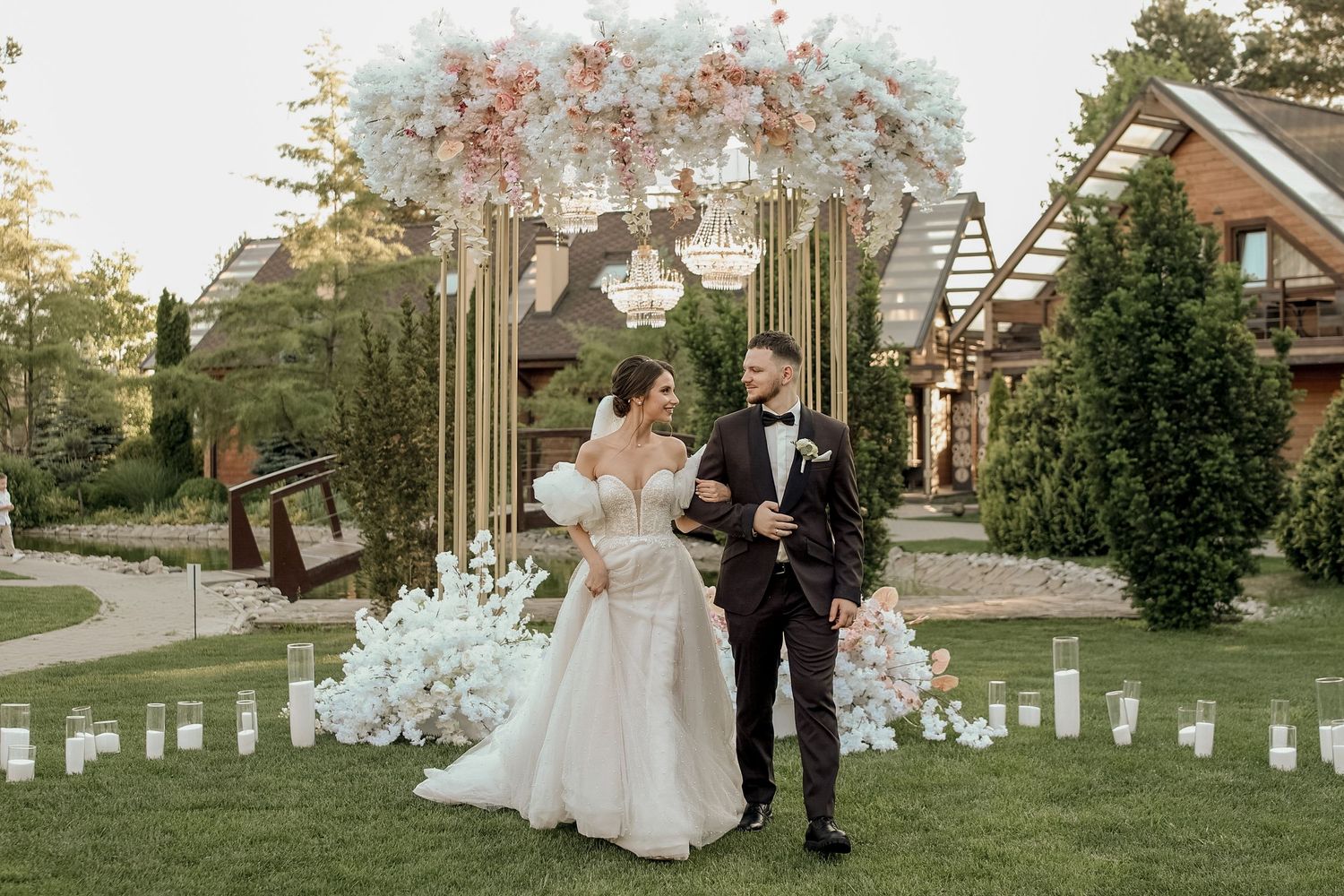 A romantic outdoor wedding ceremony setup with white floral arrangements and wooden pergola at sunset.