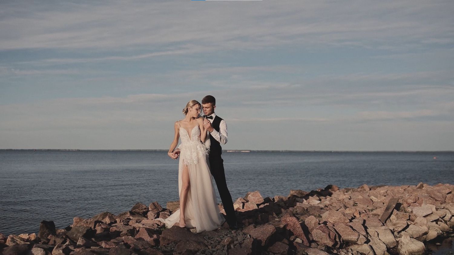 A bride and groom embrace romantically on rocky shoreline with ocean and blue sky backdrop on their wedding day.