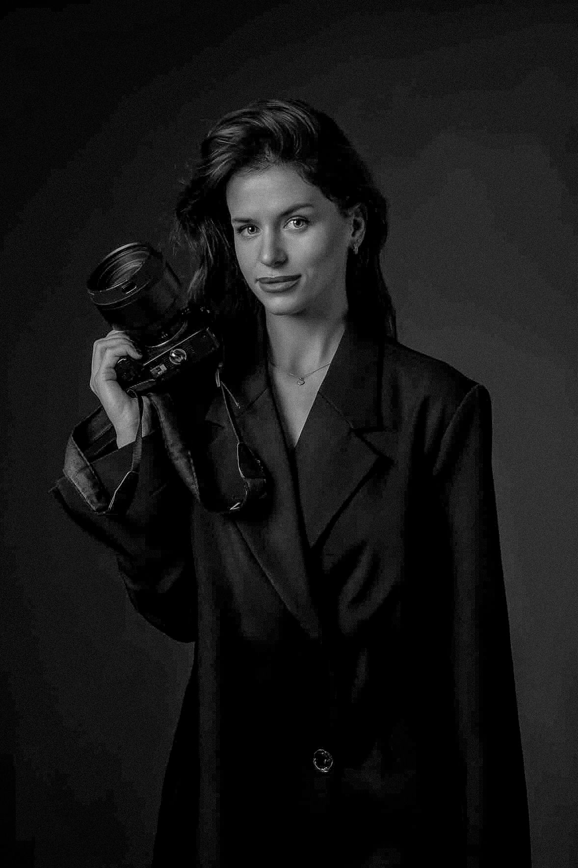 Black and white portrait of a woman in a blazer holding a camera with a telephoto lens.