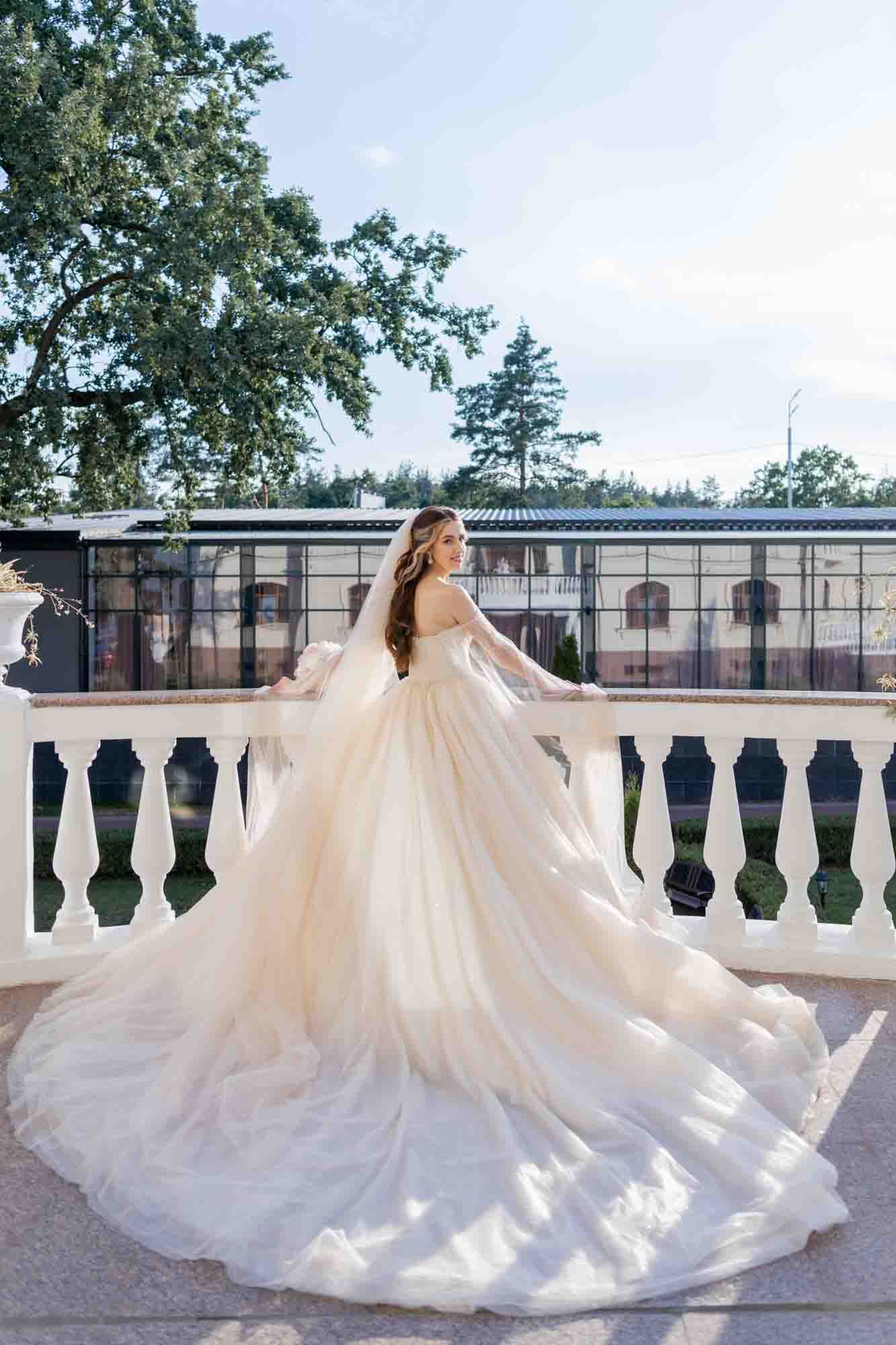 Bride in elegant champagne ballgown with long train poses on white balustrade terrace surrounded by lush trees.