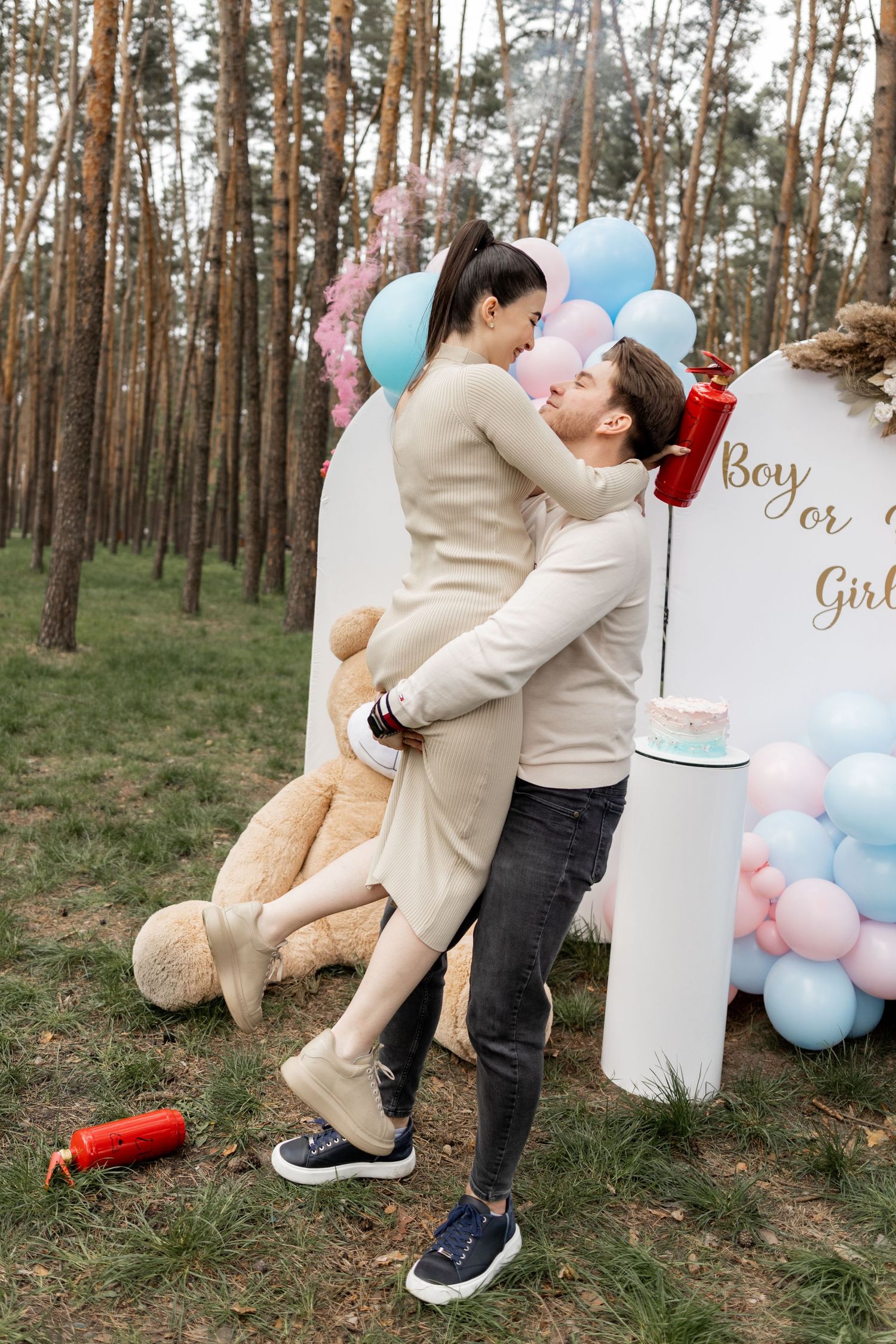 Festive gender reveal celebration in forest setting with balloon decorations and signs.