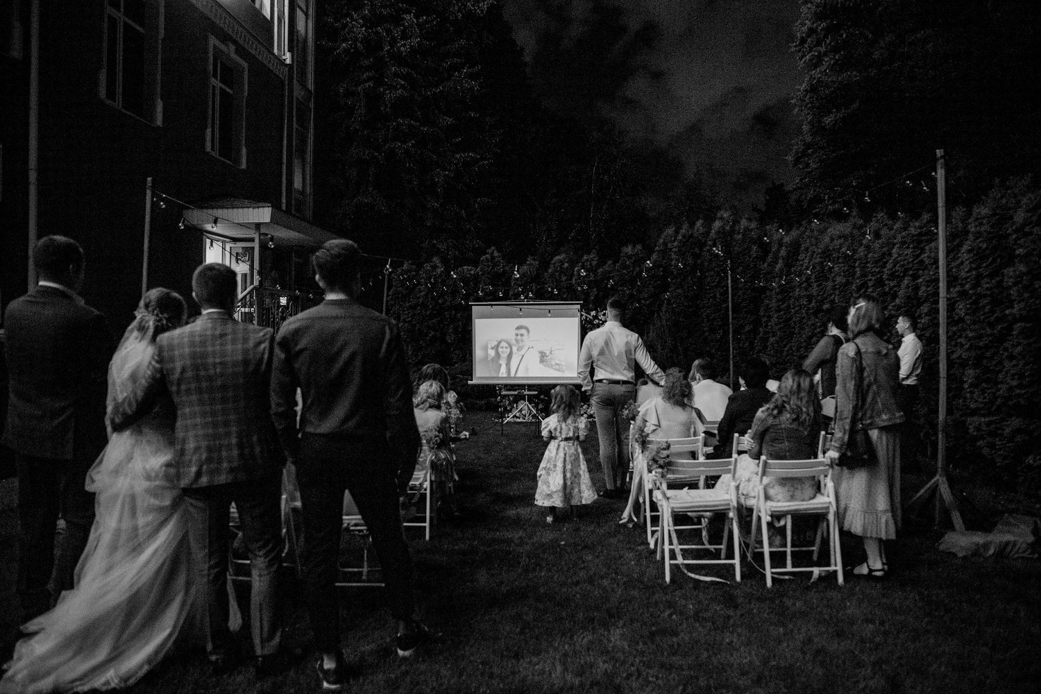 Black and white outdoor movie screening at a wedding reception with guests gathered on lawn chairs watching a projected film at dusk.