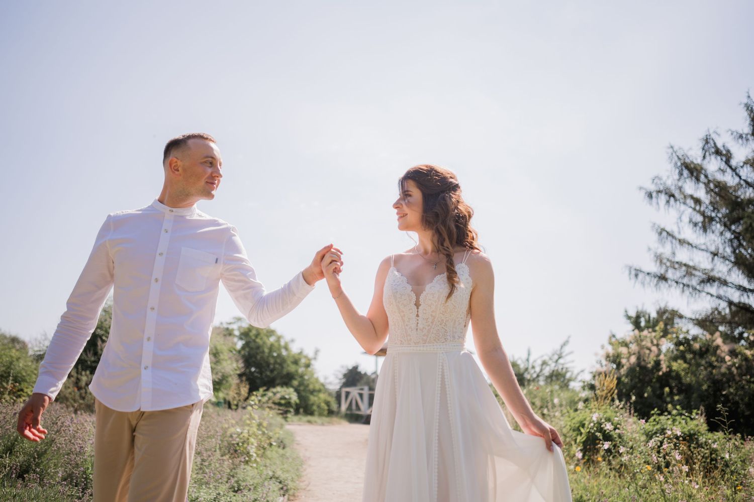 Couple in white wedding attire holding hands on a sunny outdoor path surrounded by green trees.