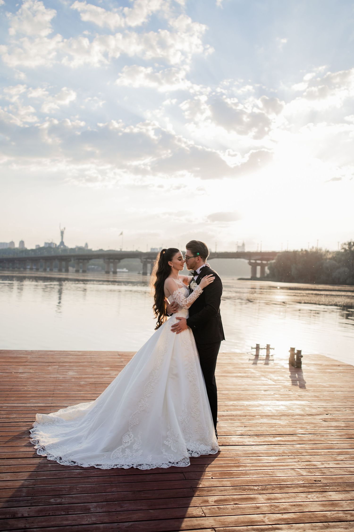 Bride and groom sharing a romantic kiss on a wooden pier by a river at sunset on their wedding day.