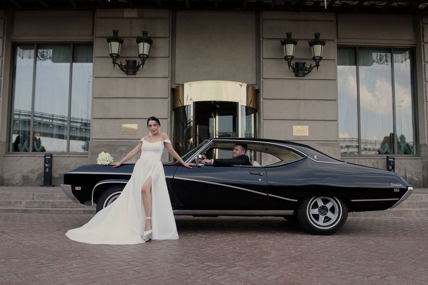 Bride in white gown poses beside a classic black muscle car outside an elegant building with a groom seated inside.
