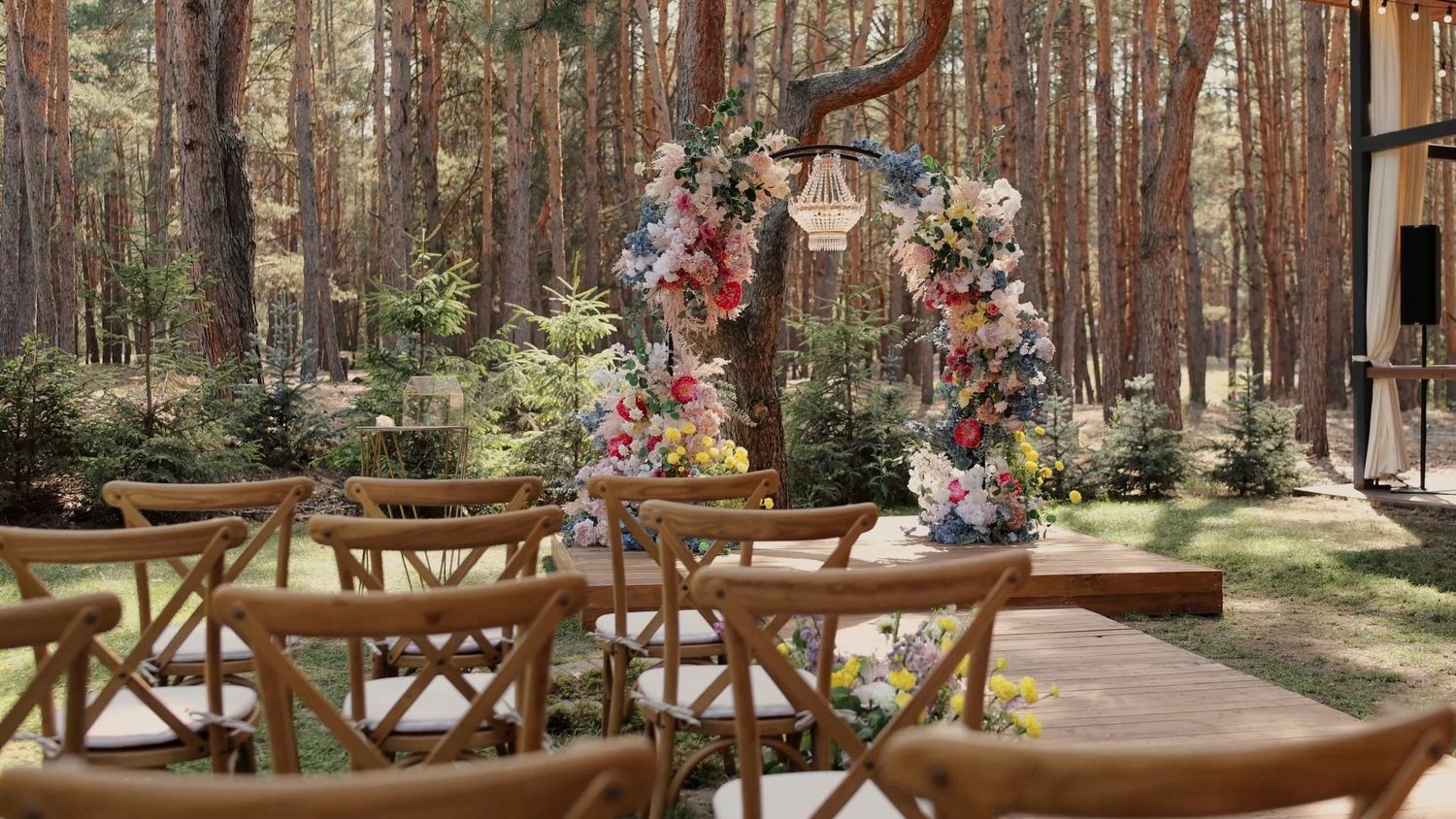 Outdoor wedding ceremony setup with wooden chairs and a floral arch in a sunlit forest.