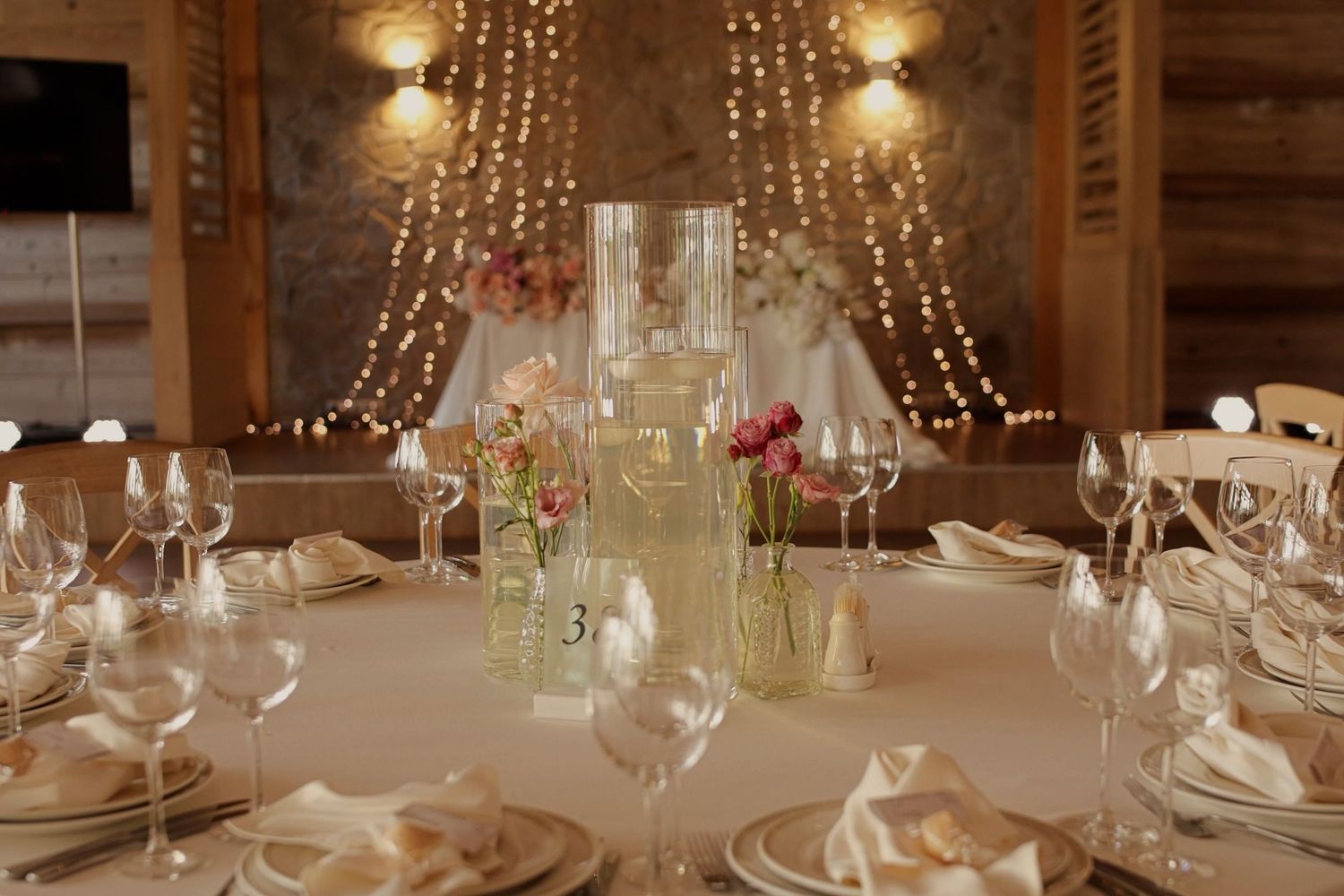 Elegant wedding reception table set with crystal glasses, floral centerpieces, and fairy lights in a warm rustic venue.