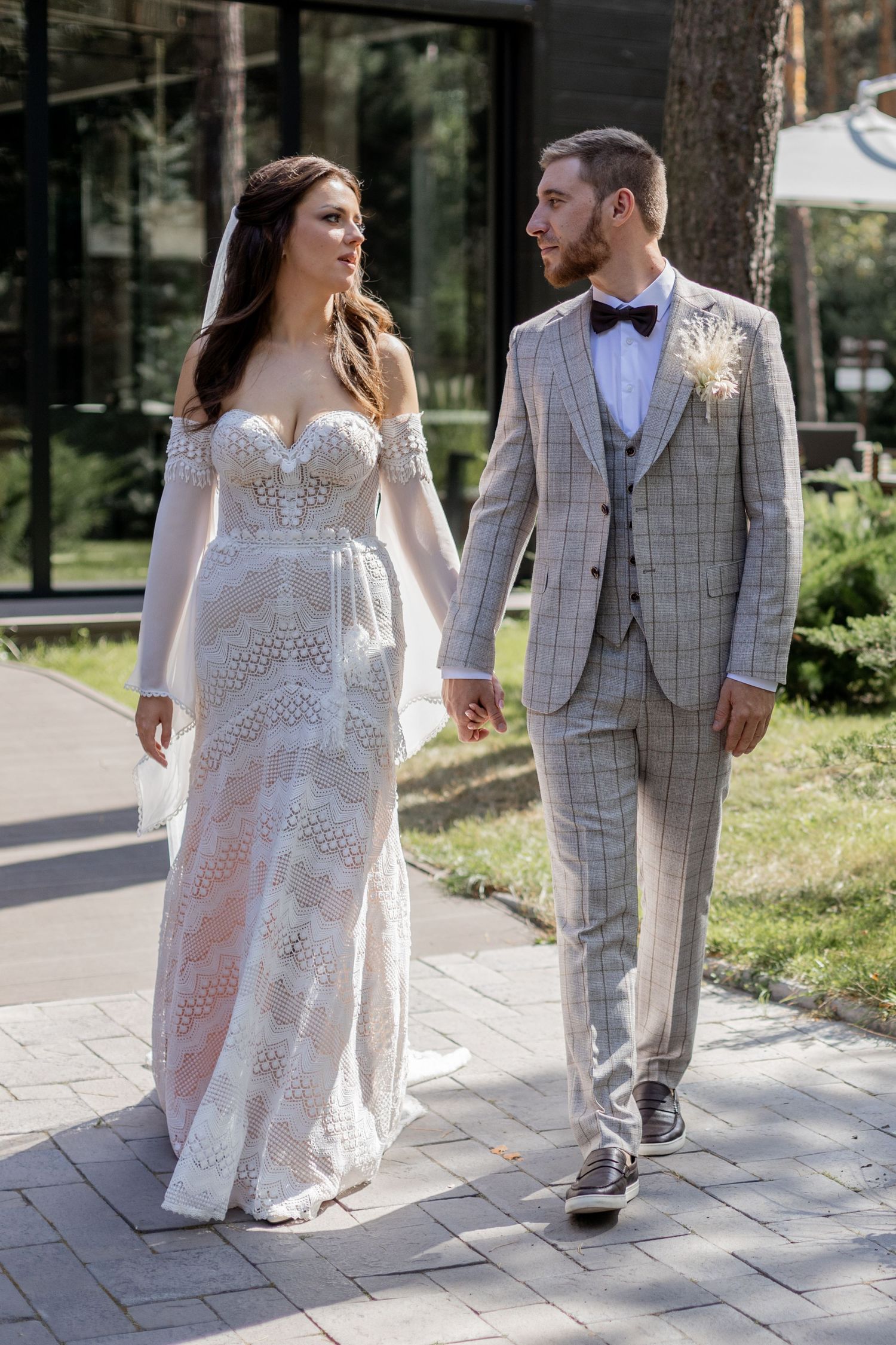 Bride in lace gown and groom in checkered suit walk together on stone pathway.