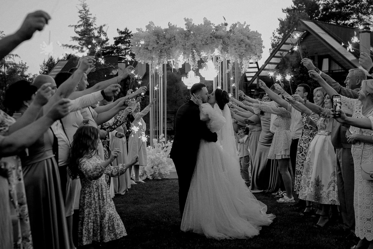 Black and white wedding photo of couple kissing during sparkler exit, surrounded by cheering guests outdoors.