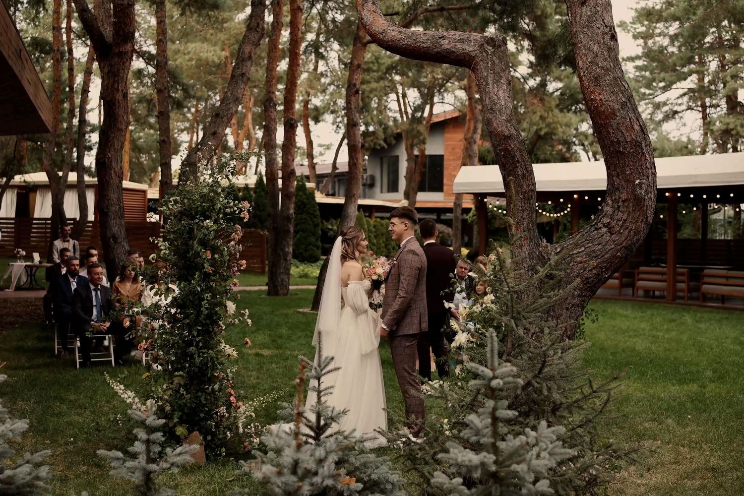 Outdoor wedding ceremony under pine trees with bride and groom at floral arch, guests seated, rustic venue backdrop.